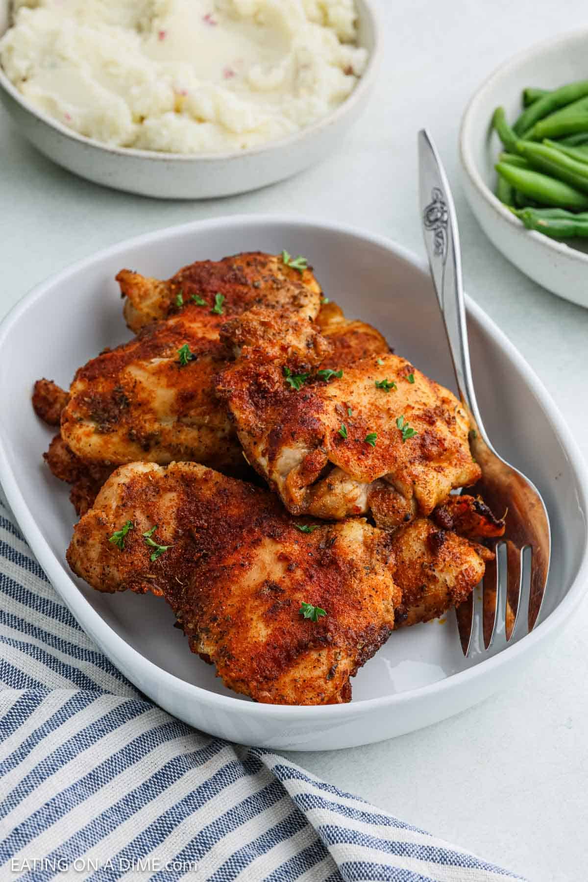 A white dish filled with seasoned, baked chicken thighs—perfectly cooked, boneless chicken thighs made in an air fryer—garnished with herbs and placed next to a fork. Mashed potatoes, green beans, and a blue-striped napkin complete the scene.