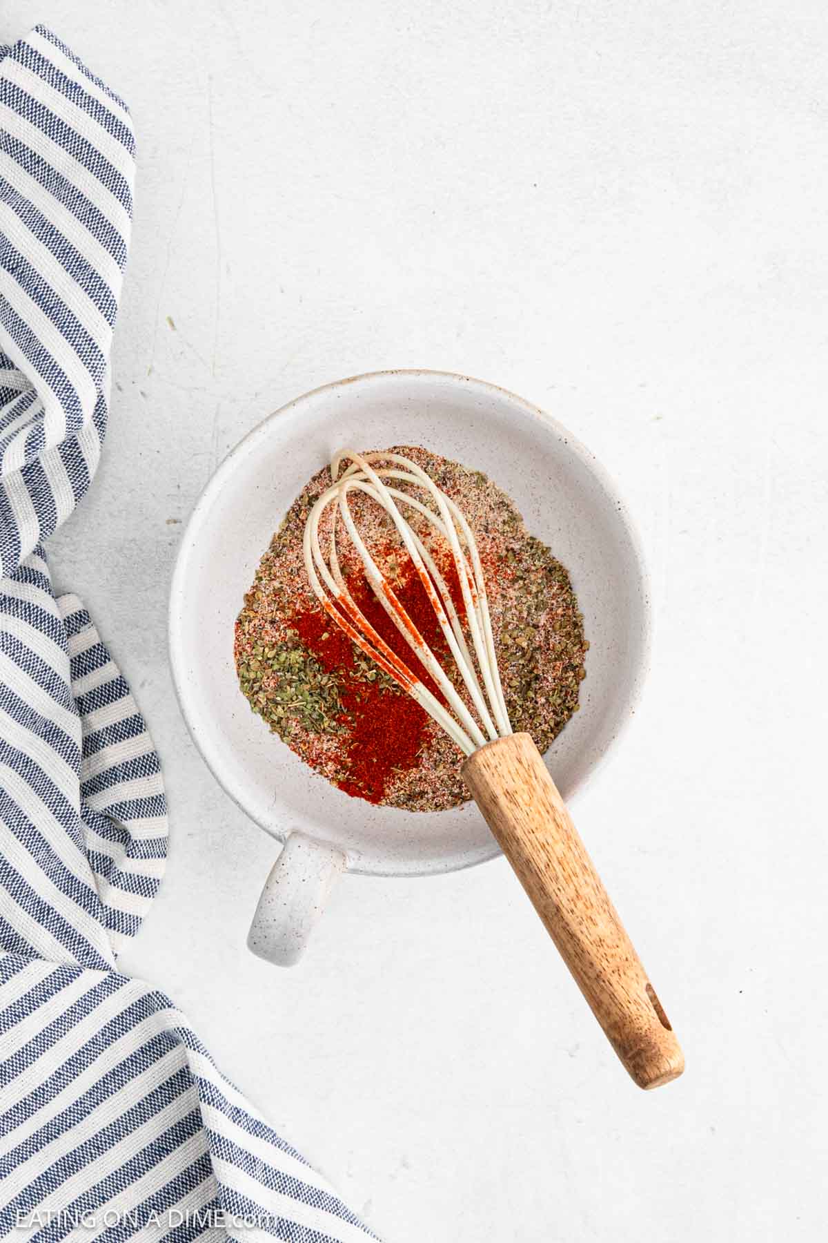 A white bowl filled with assorted ground spices and herbs, perfect for seasoning air fryer chicken, with a wooden-handled whisk resting inside. The bowl sits on a white surface next to a blue and white striped cloth.