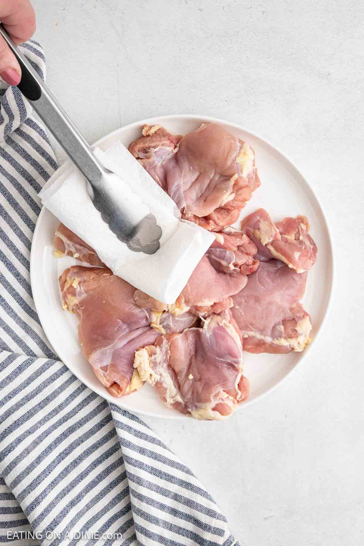 A plate of raw boneless chicken thighs is being patted dry with a paper towel held by tongs. Perfect for making tender air fryer chicken, the plate sits on a light surface next to a blue and white striped kitchen towel.