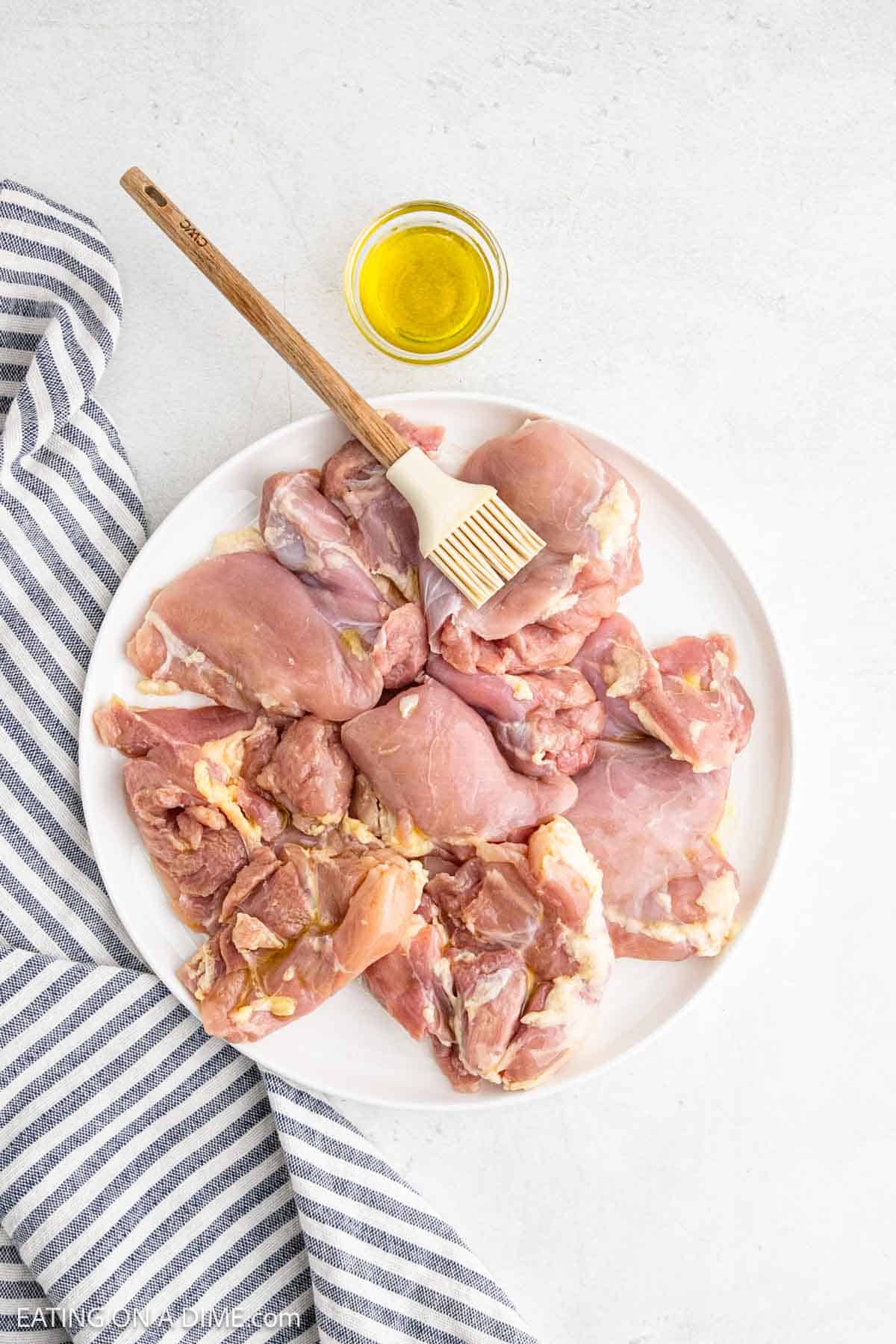 A white plate with boneless chicken thighs, a wooden-handled brush, a small bowl of olive oil, and a blue-striped cloth on a light surface—perfect prep for air fryer cooking.