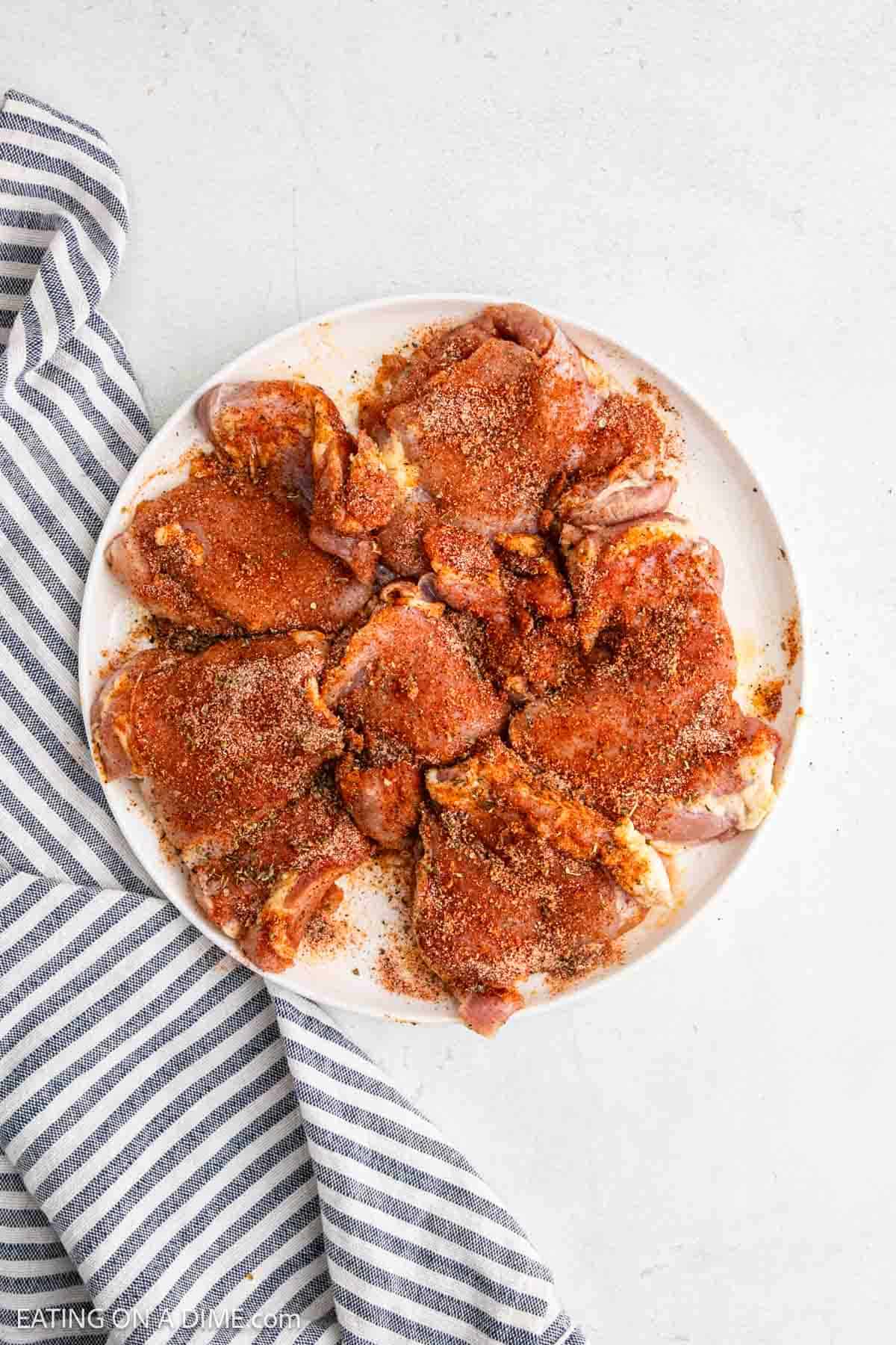 A round white plate with raw, seasoned boneless chicken thighs arranged in a circle, topped with a reddish-brown spice mix. Perfect for making air fryer chicken, the plate sits beside a folded gray and white striped cloth on a light surface.
