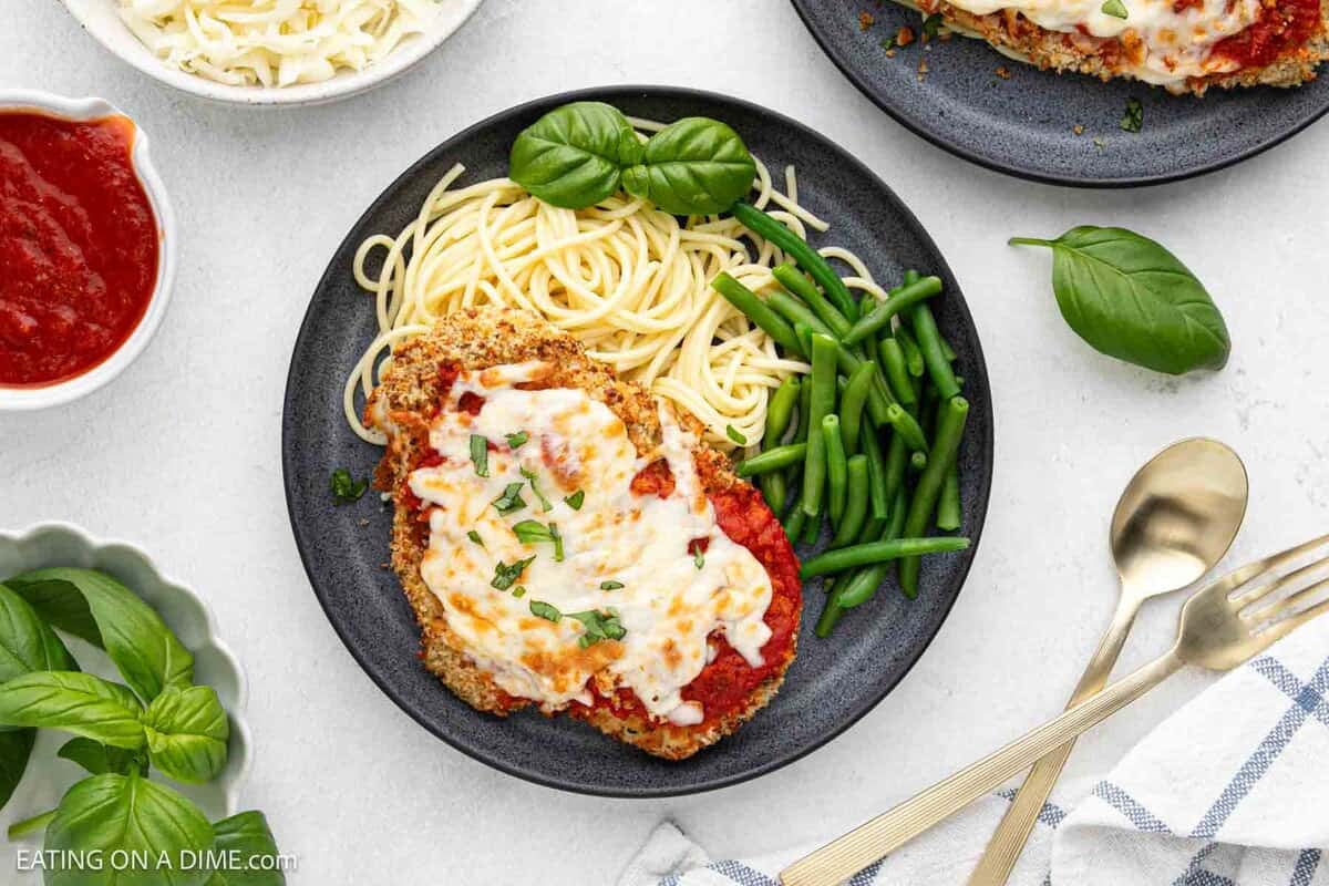A plate of Chicken Parmesan features breaded chicken topped with melted cheese and tomato sauce, served with spaghetti, green beans, and fresh basil. 