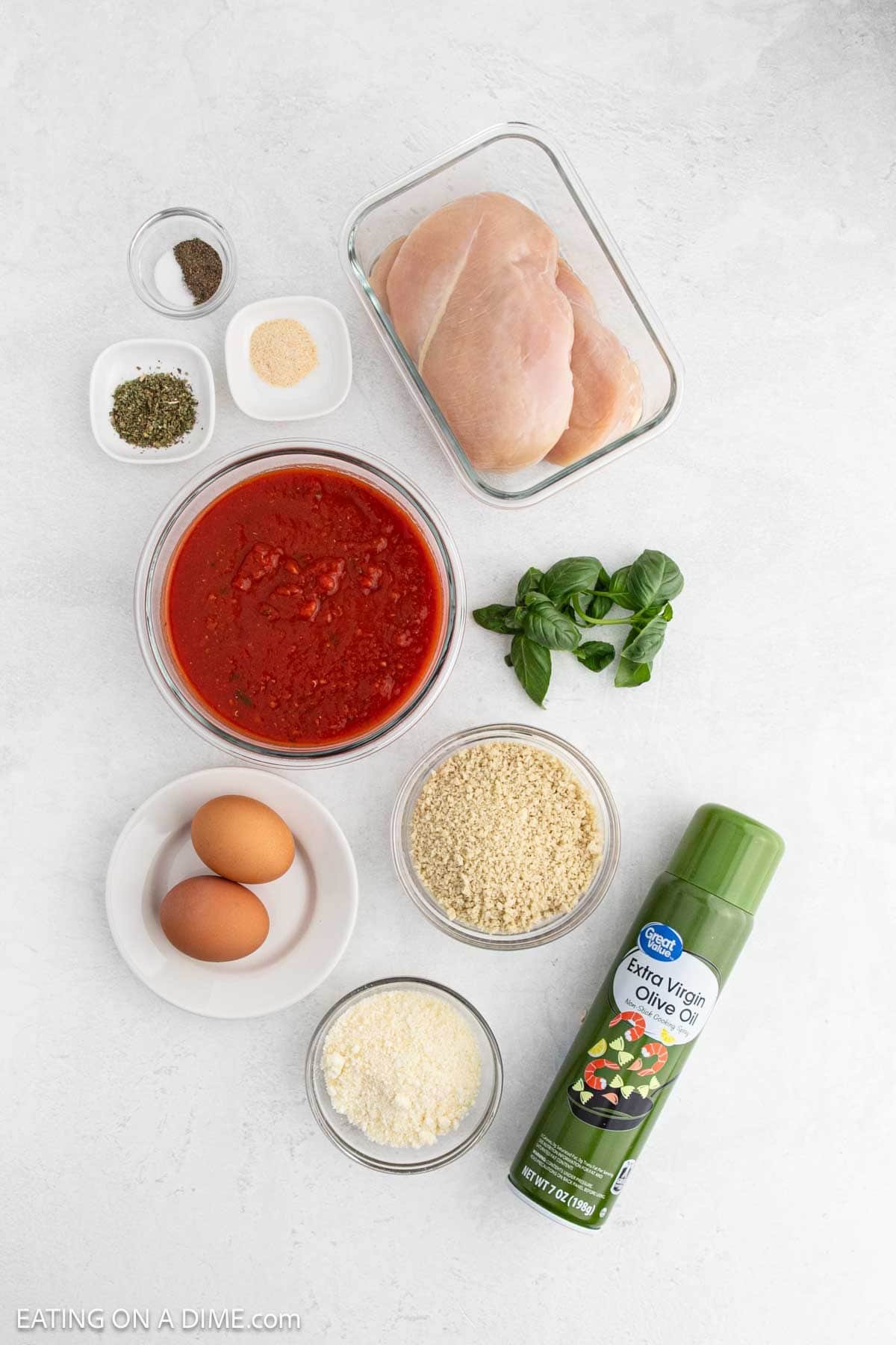 A flat lay of ingredients for Chicken Parmesan, including raw chicken breasts, canned tomatoes, eggs, breadcrumbs, grated cheese, olive oil spray—perfect for making this classic dish in your Air Fryer—plus basil and Italian seasoning on a white surface.