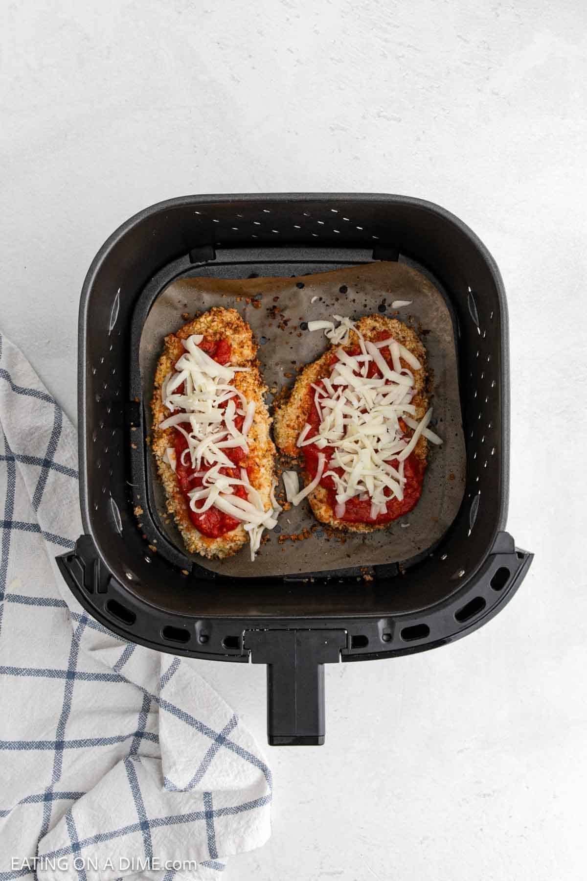 Two breaded chicken breasts for Chicken Parmesan are topped with tomato sauce and mozzarella, then placed in an air fryer basket lined with parchment paper. A white and blue checked towel sits beside the air fryer, ready for your next meal.