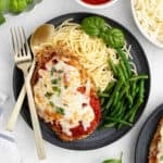 A plate of Chicken Parmesan features breaded chicken topped with melted cheese and tomato sauce, served with spaghetti, green beans, and fresh basil. Prepared in the air fryer, it’s accompanied by a fork and spoon beside the plate.