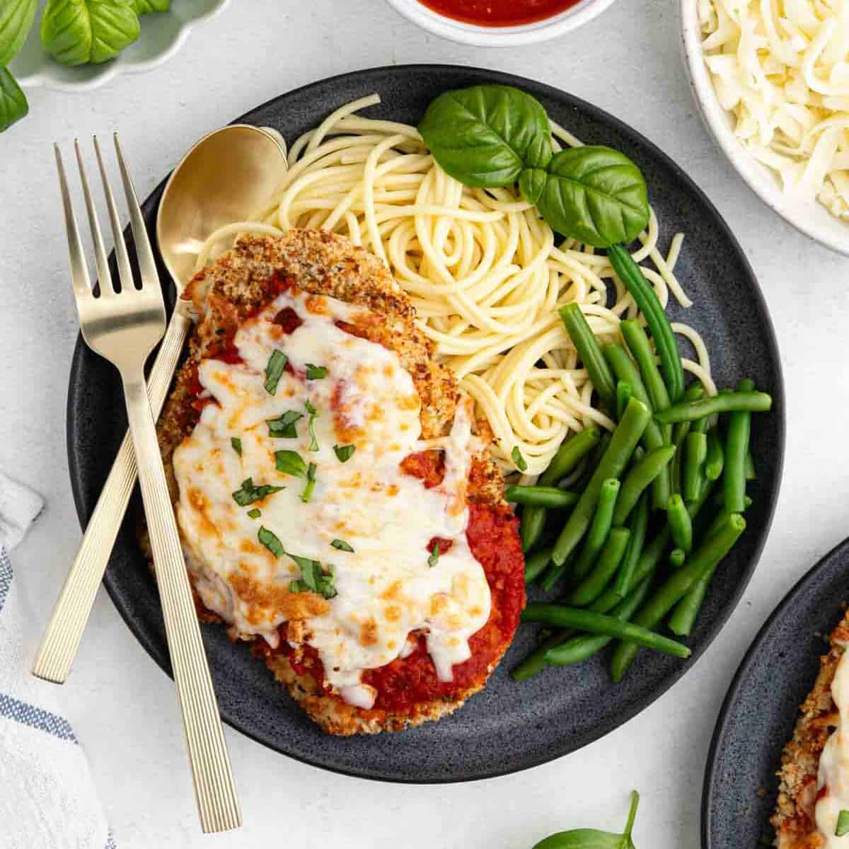 A plate of Chicken Parmesan features breaded chicken topped with melted cheese and tomato sauce, served with spaghetti, green beans, and fresh basil. Prepared in the air fryer, it’s accompanied by a fork and spoon beside the plate.