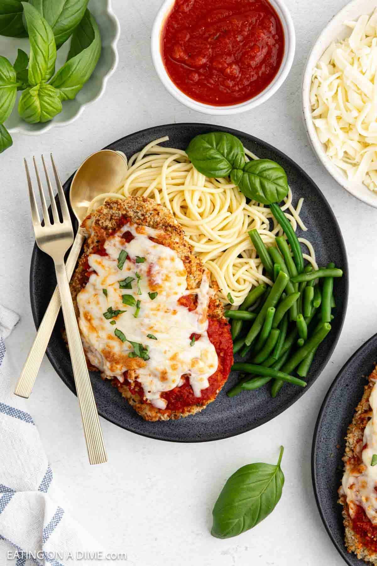 A plate with crispy Air Fryer Chicken Parmesan topped with melted cheese and tomato sauce, served alongside tender green beans and a portion of spaghetti. Fresh basil, a bowl of marinara, and shredded cheese are nearby.