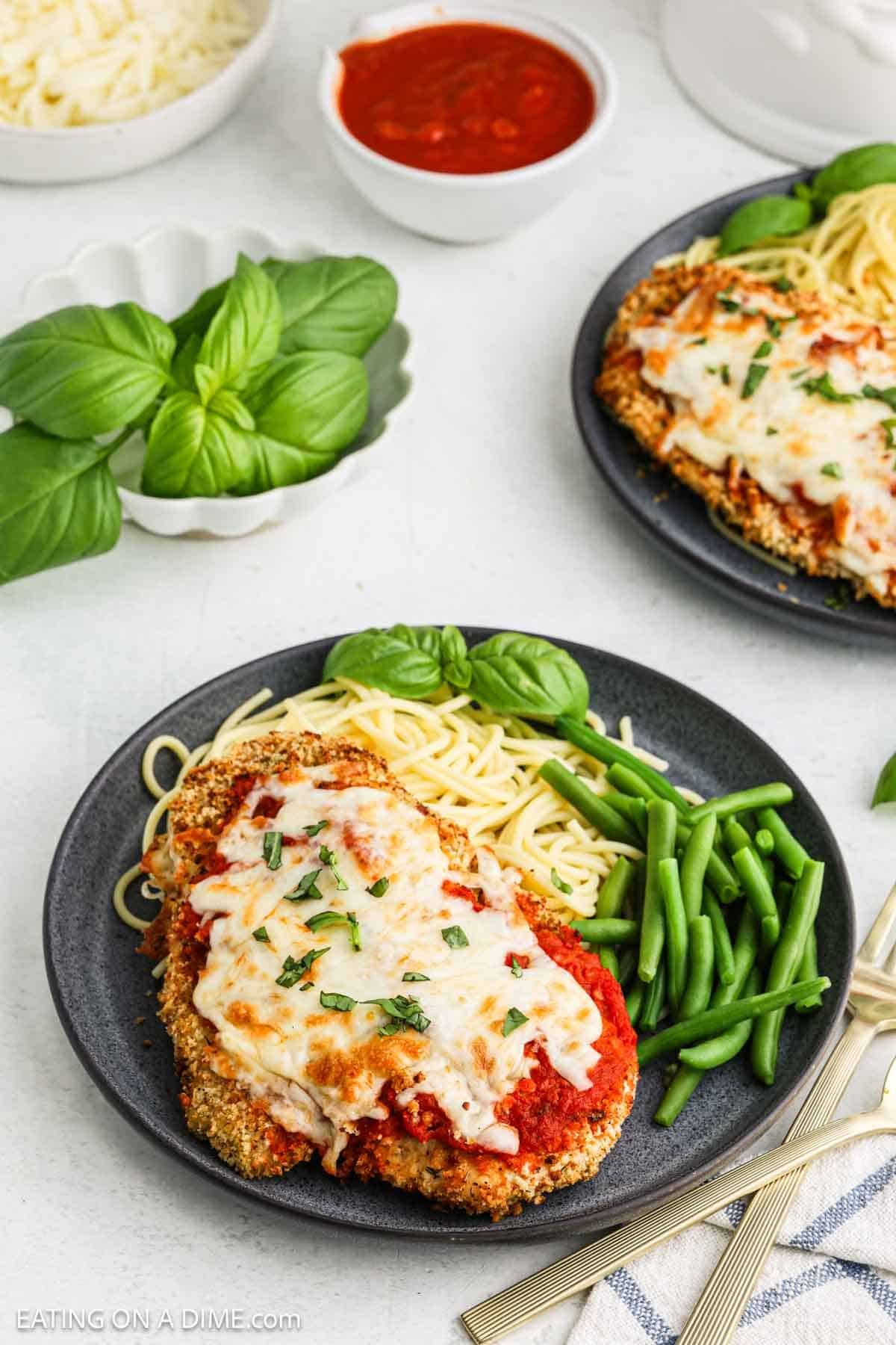 A plate of Air Fryer Chicken Parmesan topped with melted cheese and herbs, served with spaghetti, green beans, and fresh basil. Another similar plate and a bowl of marinara sauce are in the background.