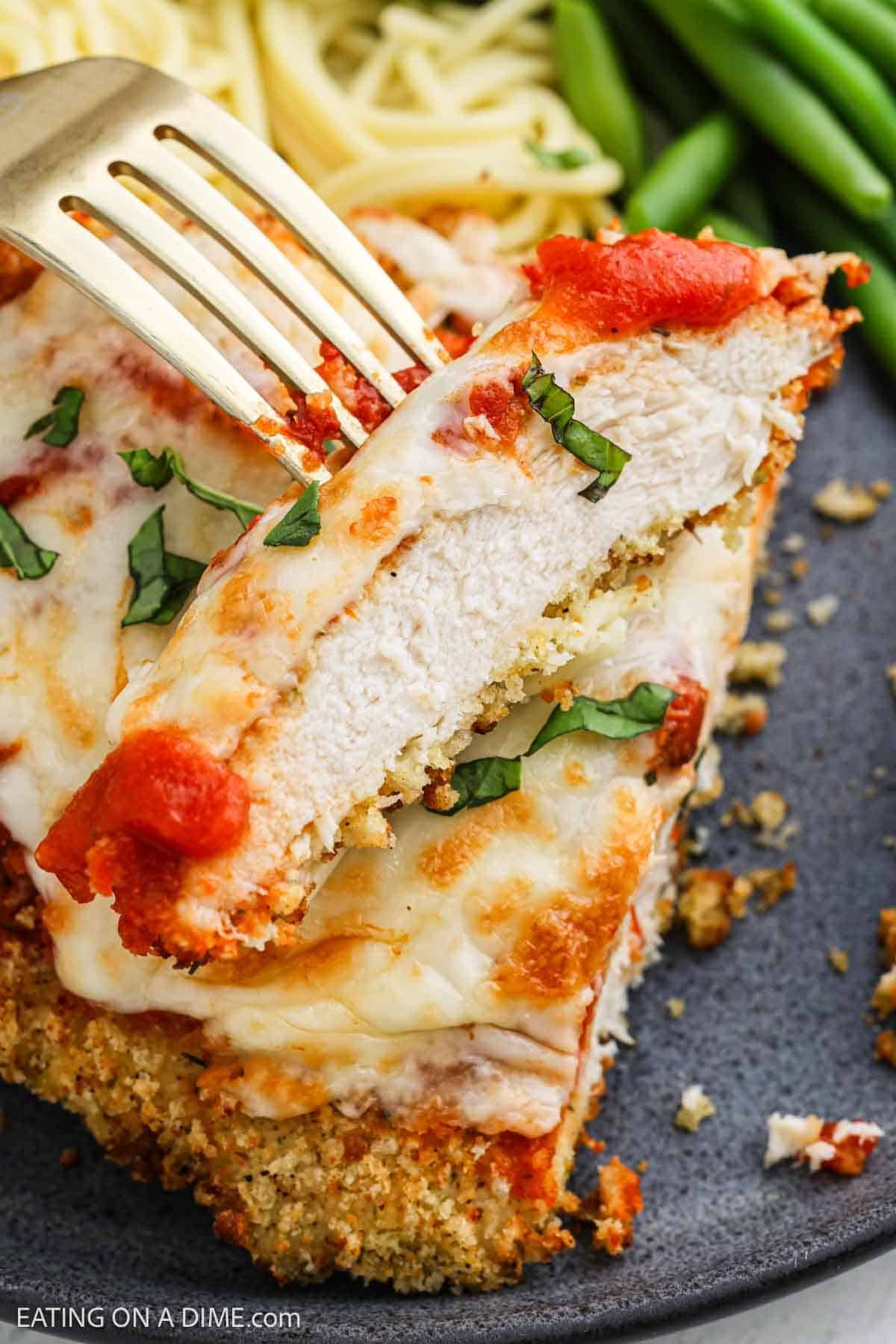 A close-up of Air Fryer Chicken Parmesan: a breaded chicken breast topped with melted cheese, marinara sauce, and fresh basil slices, being cut with a fork. Green beans and pasta are visible in the background on a dark plate.