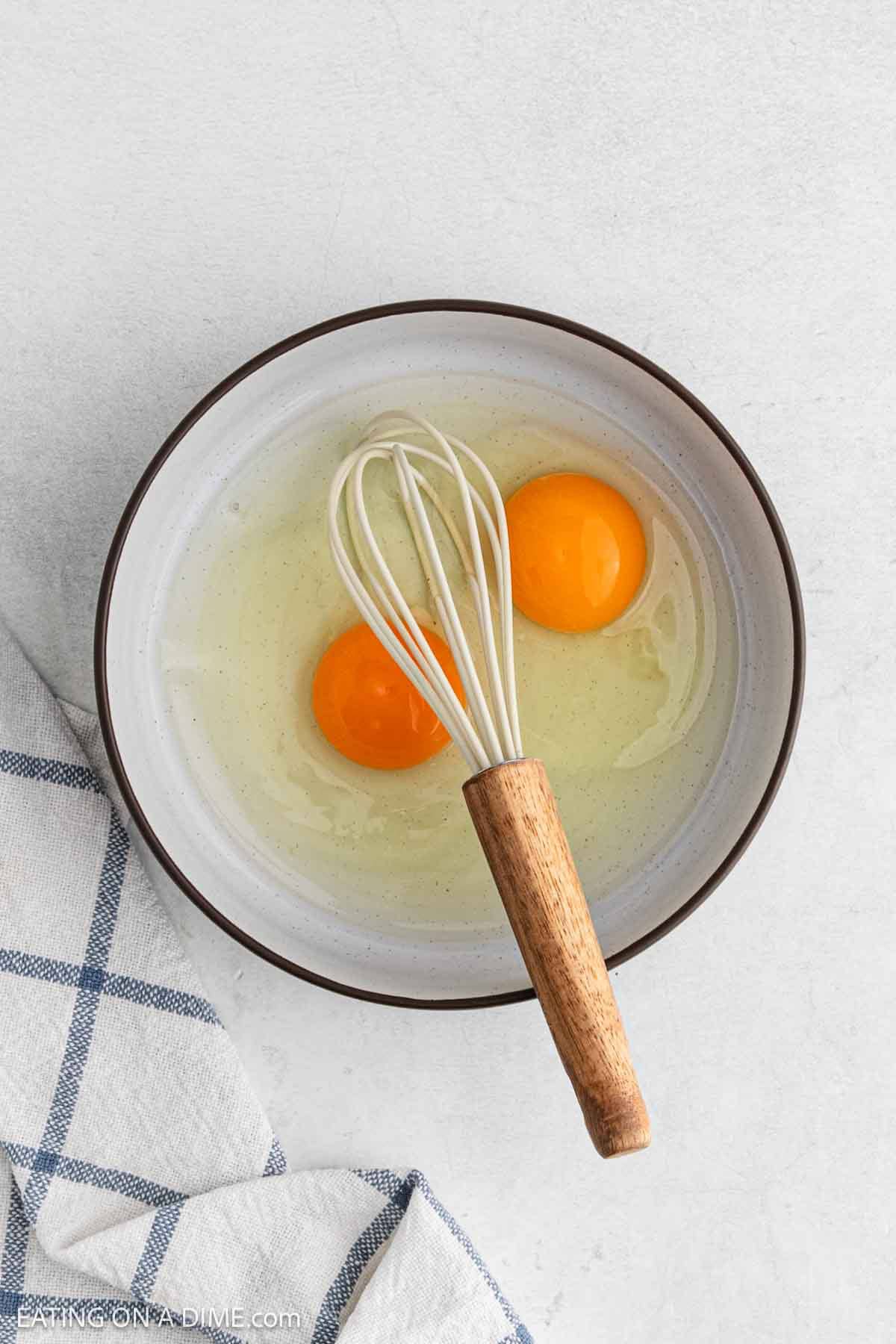 A bowl with two raw eggs and a whisk inside, placed on a light surface next to a blue and white checkered kitchen towel, ready for preparing Chicken Parmesan.