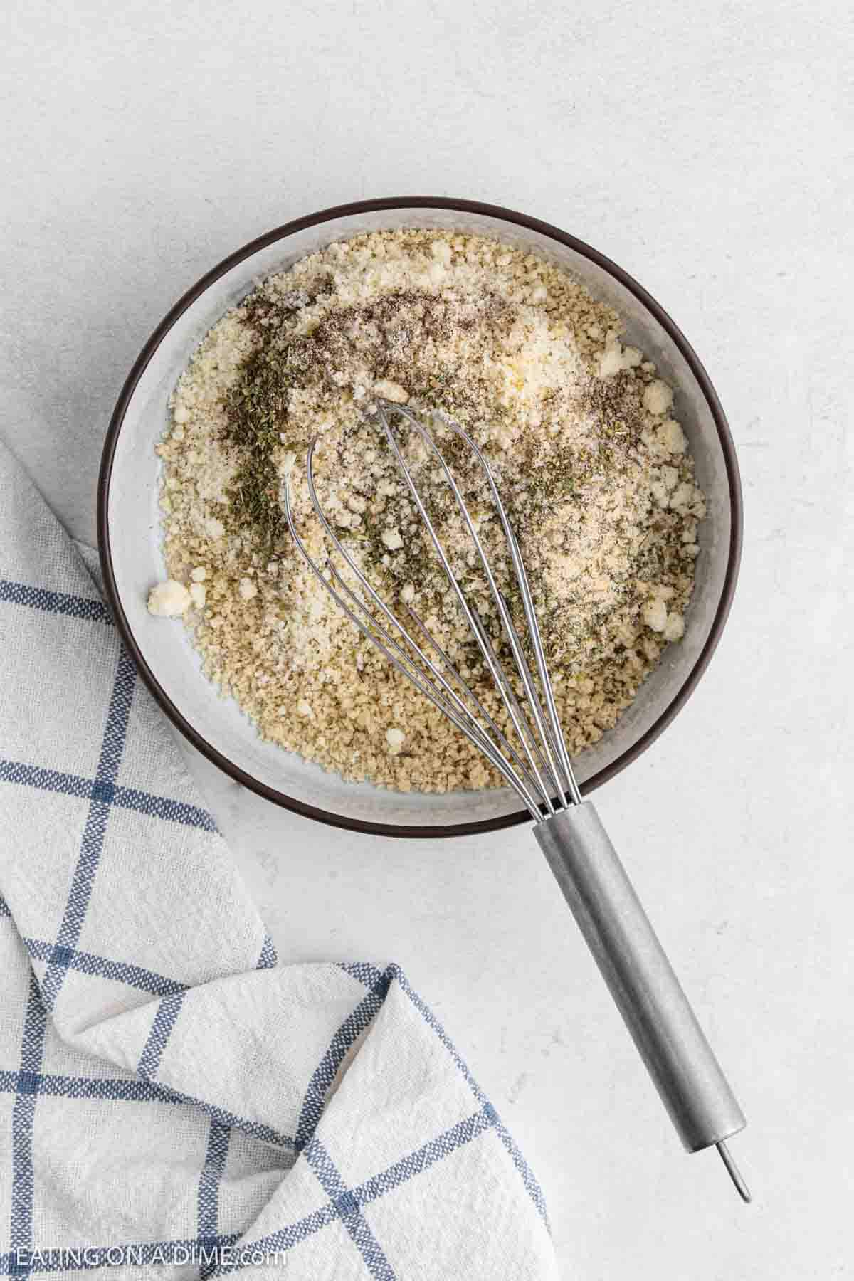 A metal whisk rests in a bowl of breadcrumbs, grated cheese, and dried herbs—perfect for coating Chicken Parmesan before cooking it in your Air Fryer. The bowl sits on a white surface next to a white towel with blue stripes.