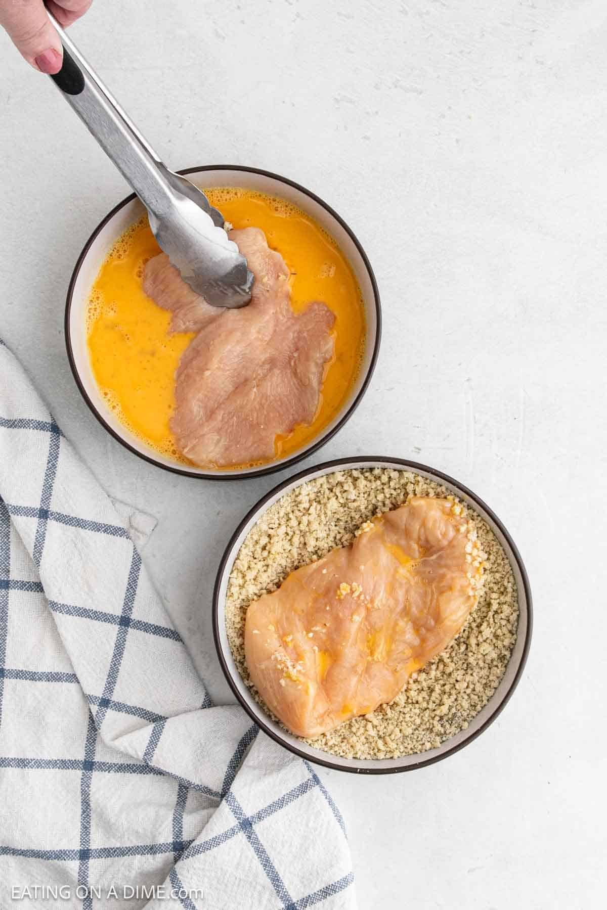 A hand uses tongs to dip raw chicken breast into a bowl of beaten eggs, with another bowl of breadcrumbs nearby—perfect prep for Chicken Parmesan or crispy Air Fryer Chicken, with a checkered kitchen towel on the side.