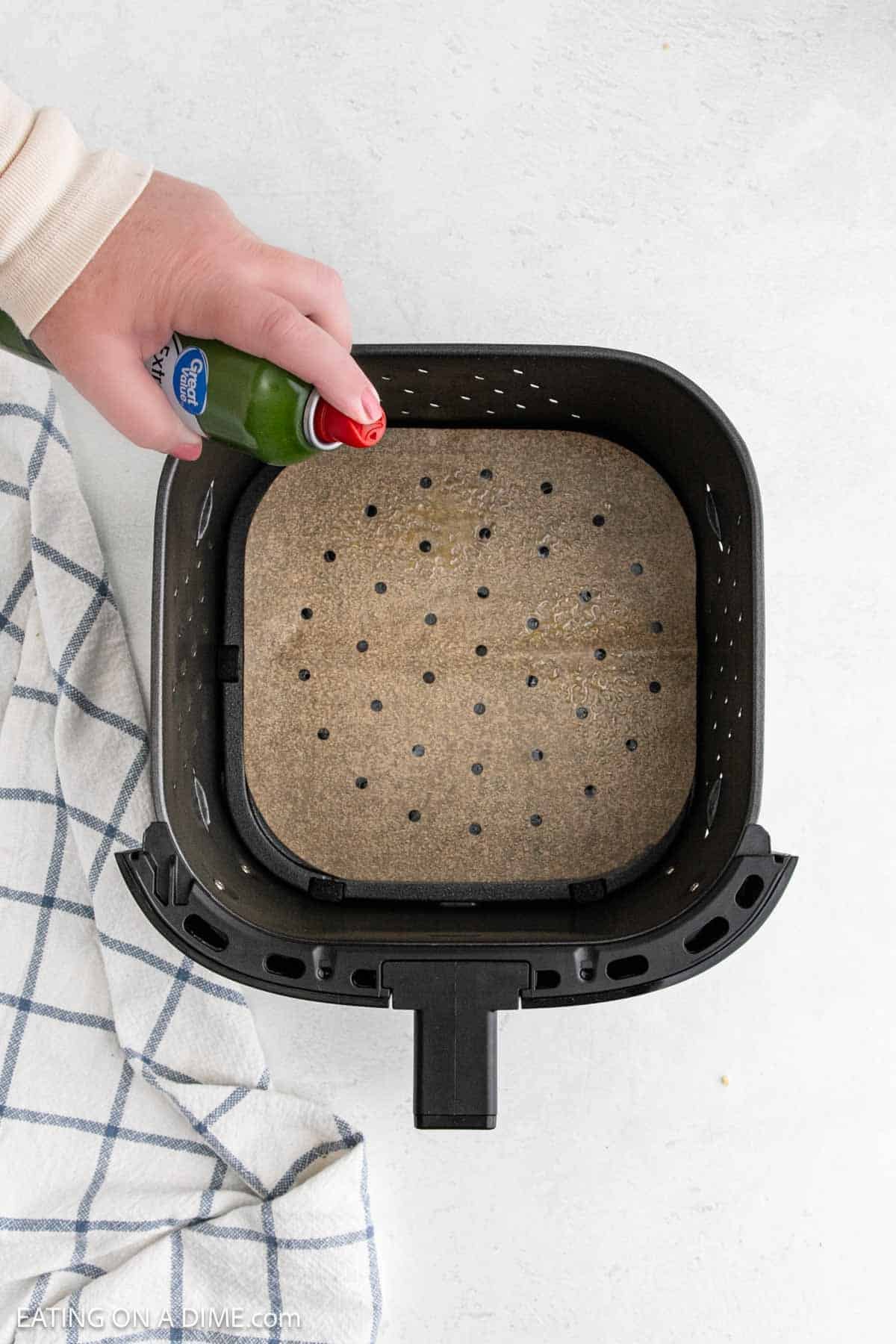 A hand sprays nonstick cooking spray into an air fryer basket lined with perforated parchment paper, prepping it for a delicious Chicken Parmesan. A striped cloth rests nearby on a white surface.