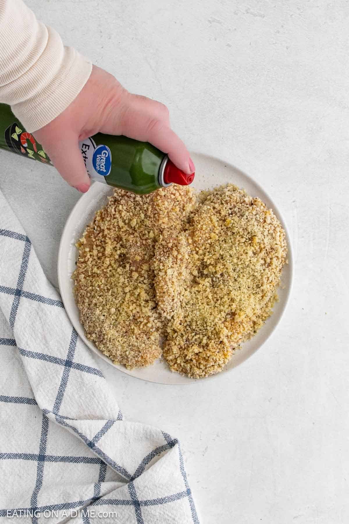 A hand sprays cooking oil over two breaded chicken cutlets, perfect for making Chicken Parmesan in the air fryer, on a white plate with a blue and white checkered towel nearby on a light surface.