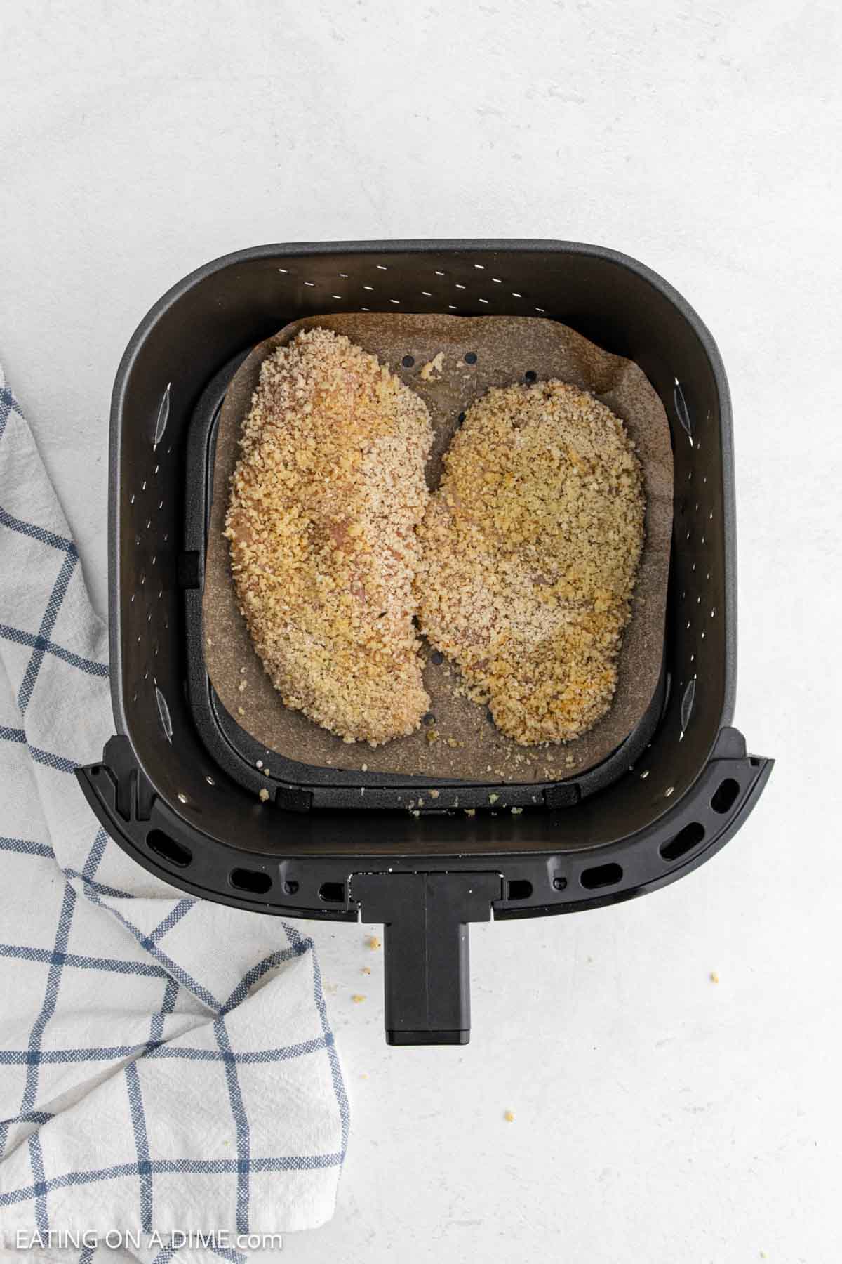 An air fryer basket contains two crispy Chicken Parmesan breasts on parchment paper. A white and blue checkered towel is partially visible beside the basket on a white surface.
