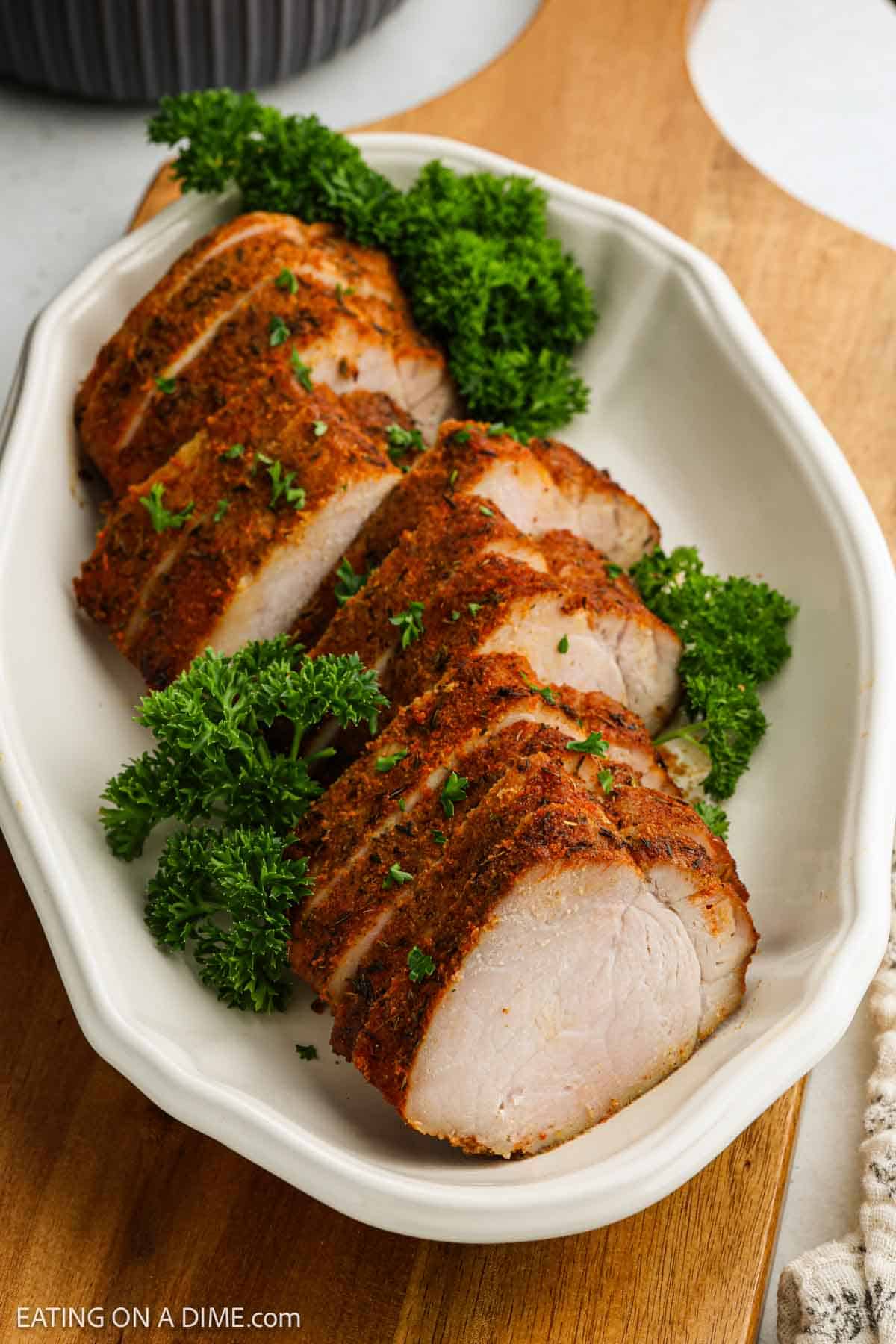 Air fryer pork tenderloin with a seasoned crust, sliced and arranged on a white platter, then garnished with fresh parsley.