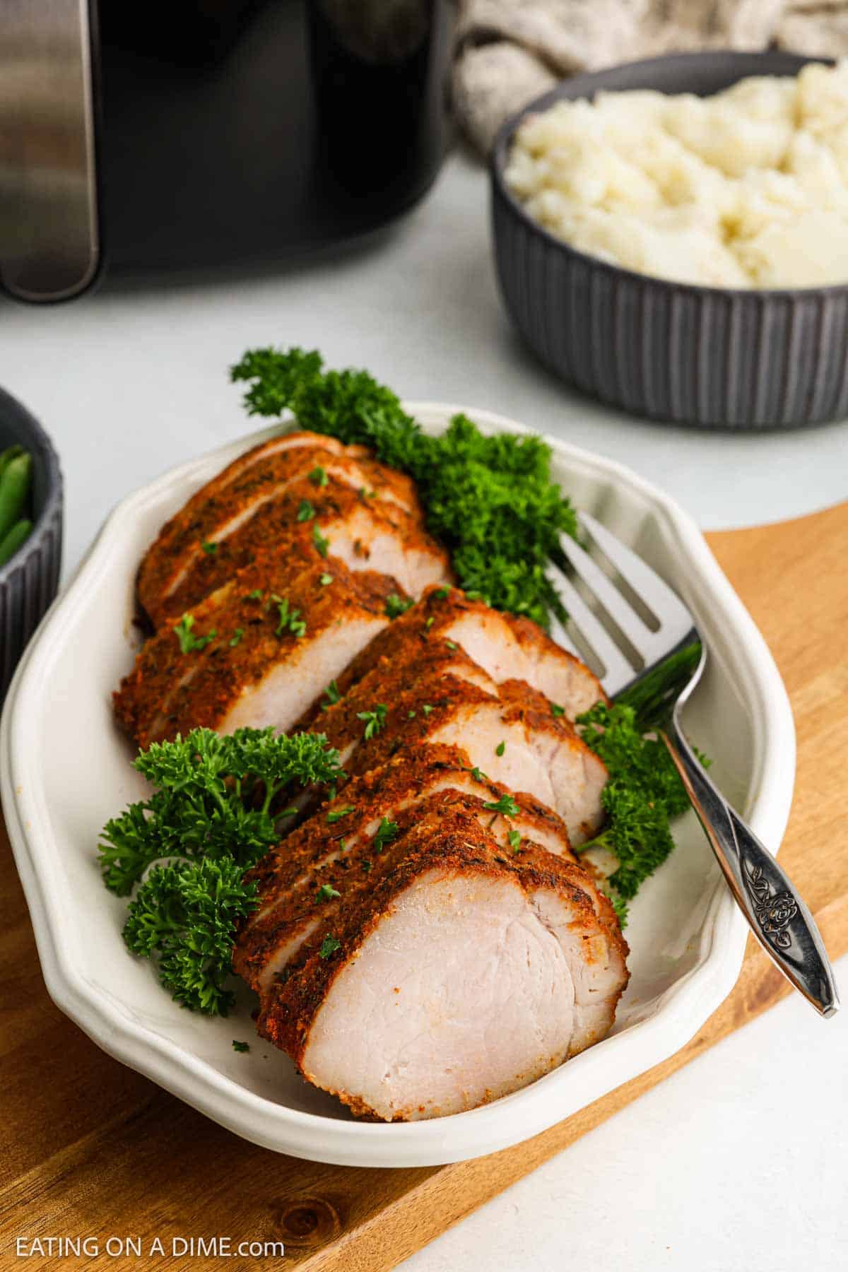 Sliced, seasoned Air Fryer Pork Tenderloin garnished with parsley on a white serving dish with a fork, set on a wooden board. A bowl of mashed potatoes is in the background.