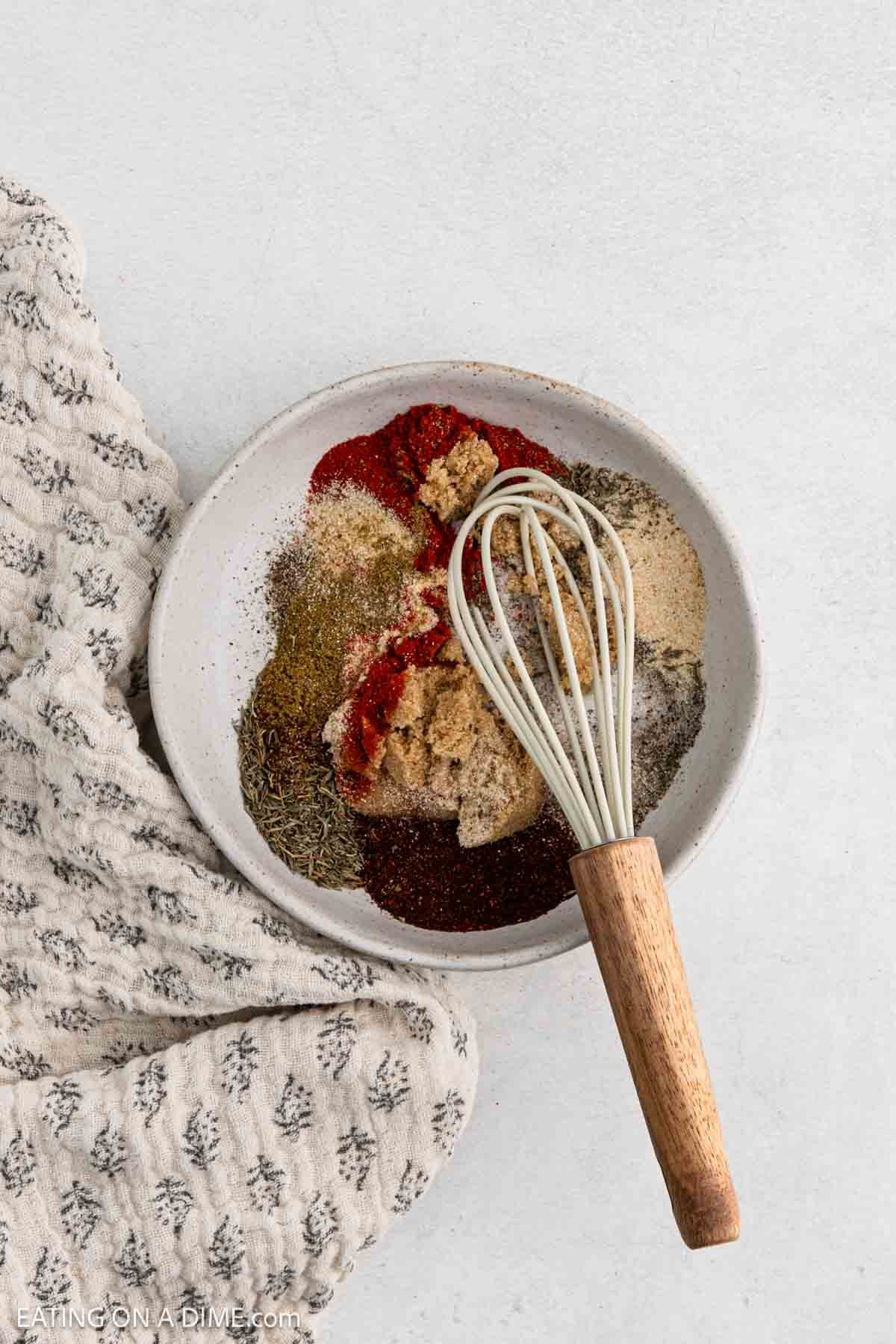 A bowl filled with various spices and brown sugar for pork tenderloin sits next to a patterned cloth. A small whisk with a wooden handle rests inside the bowl, perfect for mixing up an air fryer seasoning blend.
