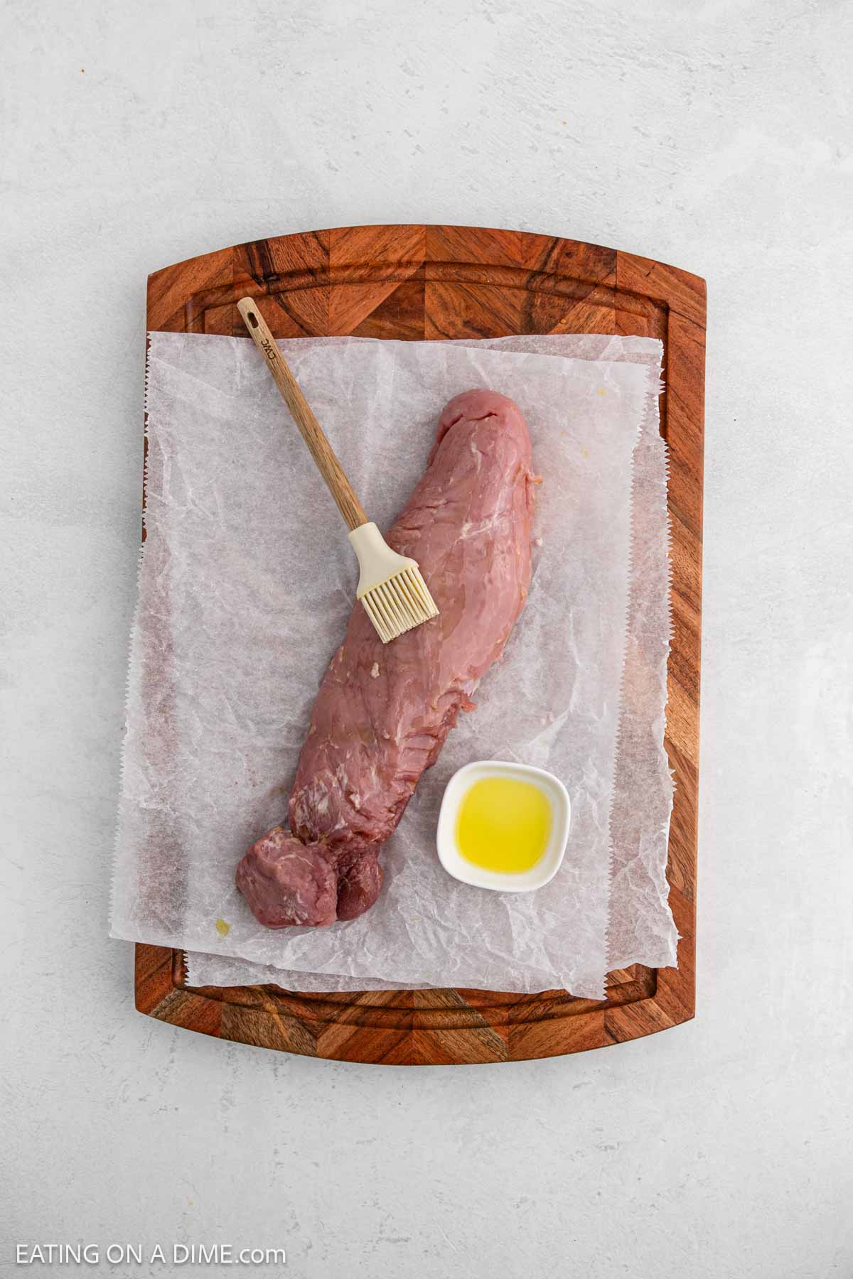 Raw pork tenderloin on parchment paper placed on a wooden cutting board, with a small bowl of oil and a basting brush resting on top—perfectly prepped for delicious air fryer pork tenderloin recipes.