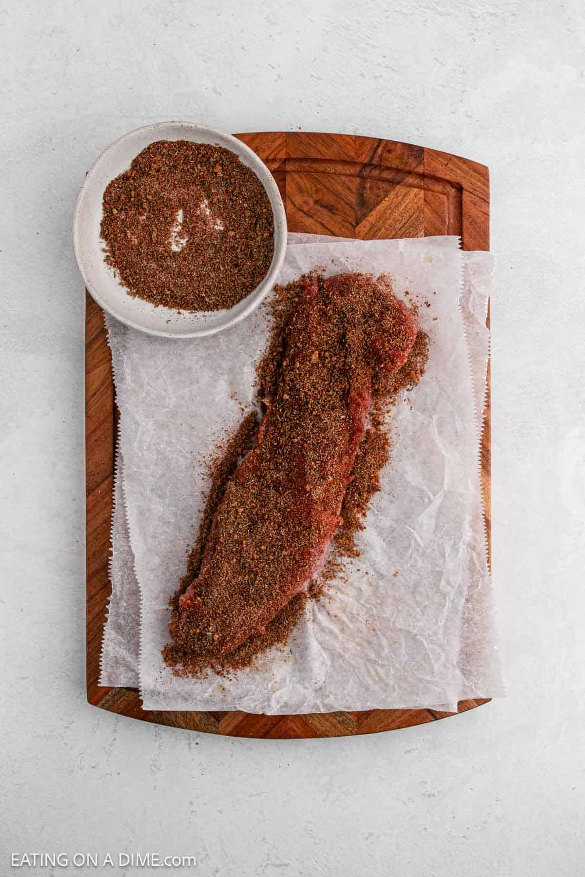 A raw pork tenderloin coated with dry seasoning rub rests on parchment paper atop a wooden cutting board. A bowl of extra seasoning is placed nearby, ready for cooking in the air fryer.