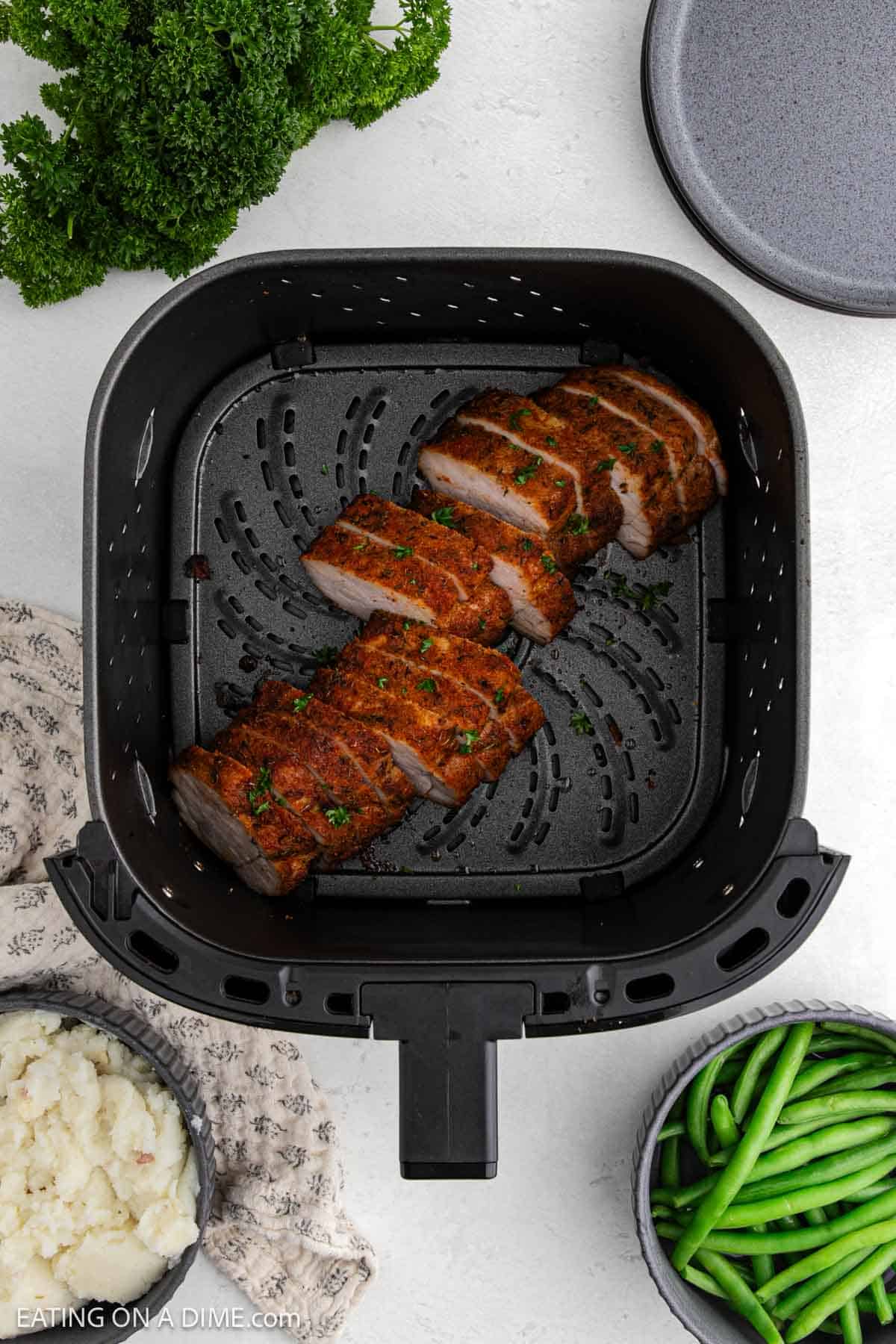 Air Fryer Pork Tenderloin sliced and seasoned in an air fryer basket, surrounded by bowls of mashed potatoes, green beans, fresh parsley, and a pot lid on a light countertop.