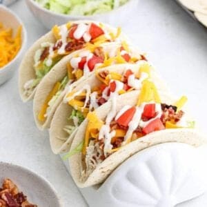 Three soft tacos filled with shredded chicken, lettuce, cheddar cheese, diced tomatoes, bacon bits, and ranch dressing, held upright in a white taco holder. Perfect for Meal Plan 54. Bowls of toppings are in the background.