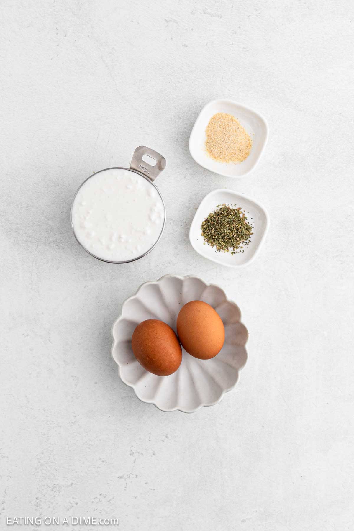 A measuring cup of milk, a small dish of dried herbs, a small dish of garlic powder, and two eggs set out for making Cottage Cheese Flatbread are arranged on a white surface.