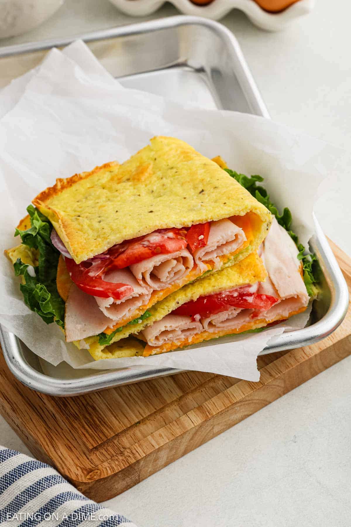 A sandwich made with folded, golden cottage cheese flatbread instead of bread, filled with lettuce, tomato, turkey, and cheese, is served on parchment paper in a metal tray on a wooden board.