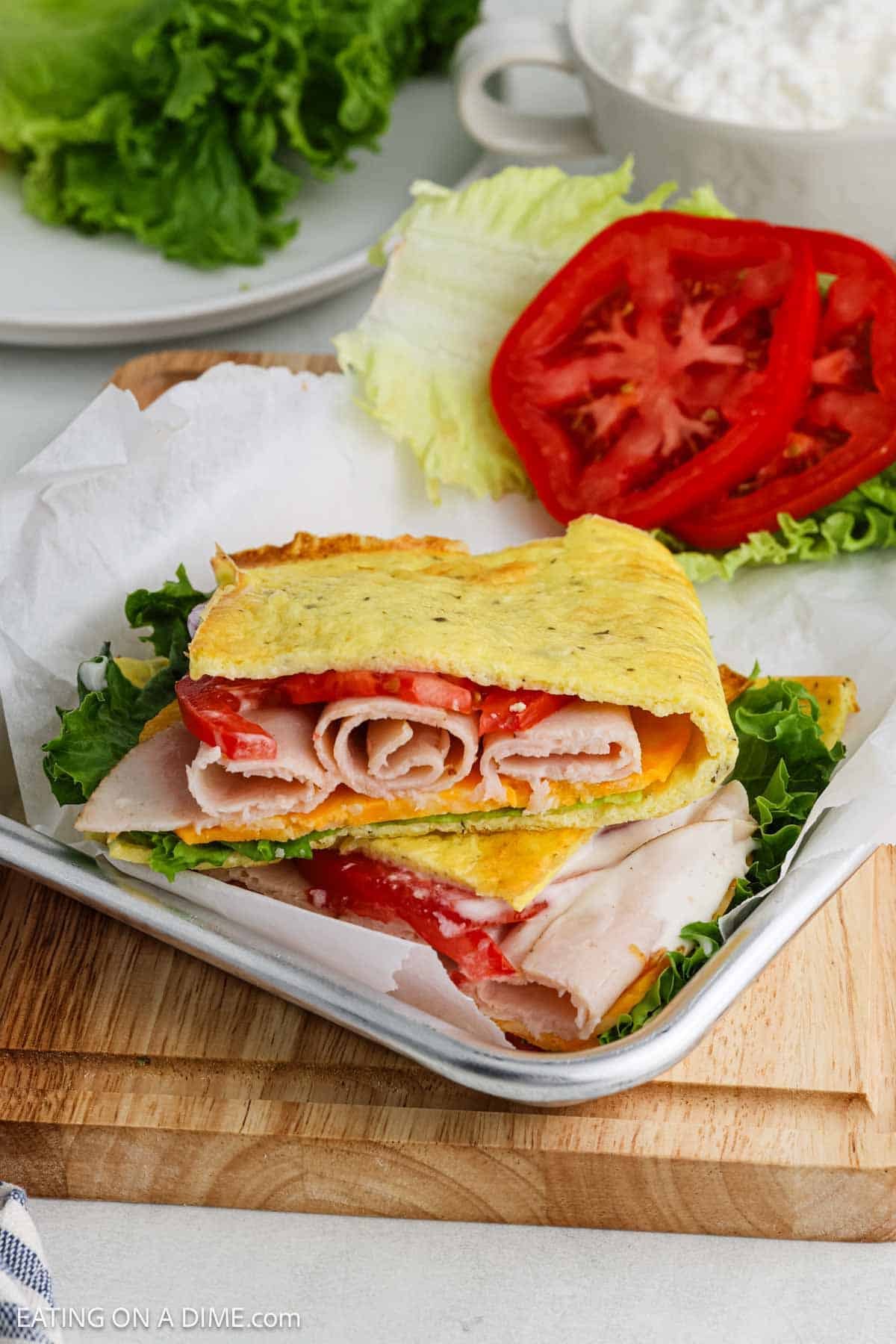 A sandwich with sliced turkey, lettuce, and tomato is wrapped in a folded cottage cheese flatbread instead of bread, served on a tray with extra lettuce and tomato slices on the side.
