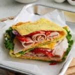 A sandwich with folded deli turkey, lettuce, tomato, and onion, served between two pieces of golden cottage cheese flatbread on parchment paper and a metal tray.
