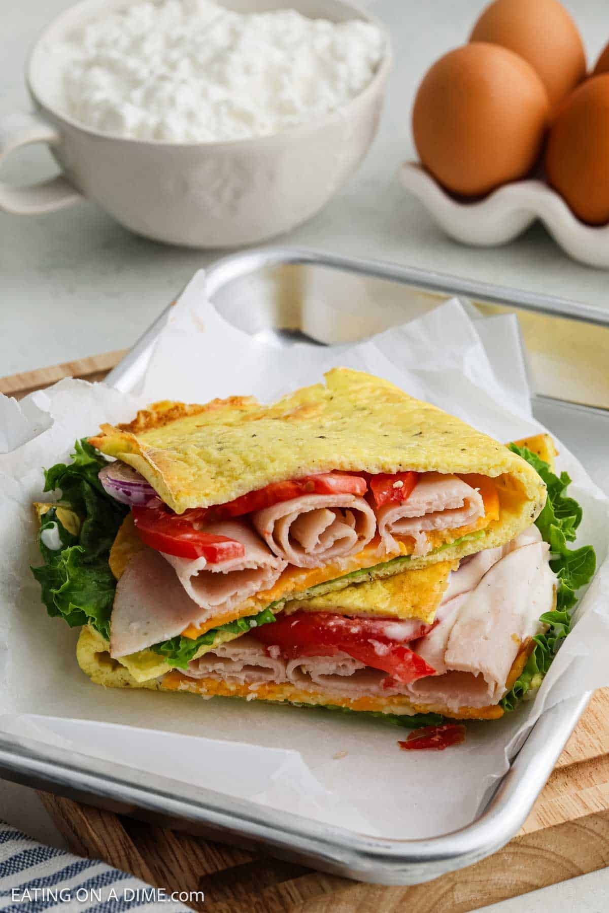 A folded sandwich with sliced turkey, lettuce, tomato, and red onions is wrapped in soft flatbread and sits on parchment paper. In the background, a bowl of creamy cottage cheese and a group of brown eggs complete the scene.