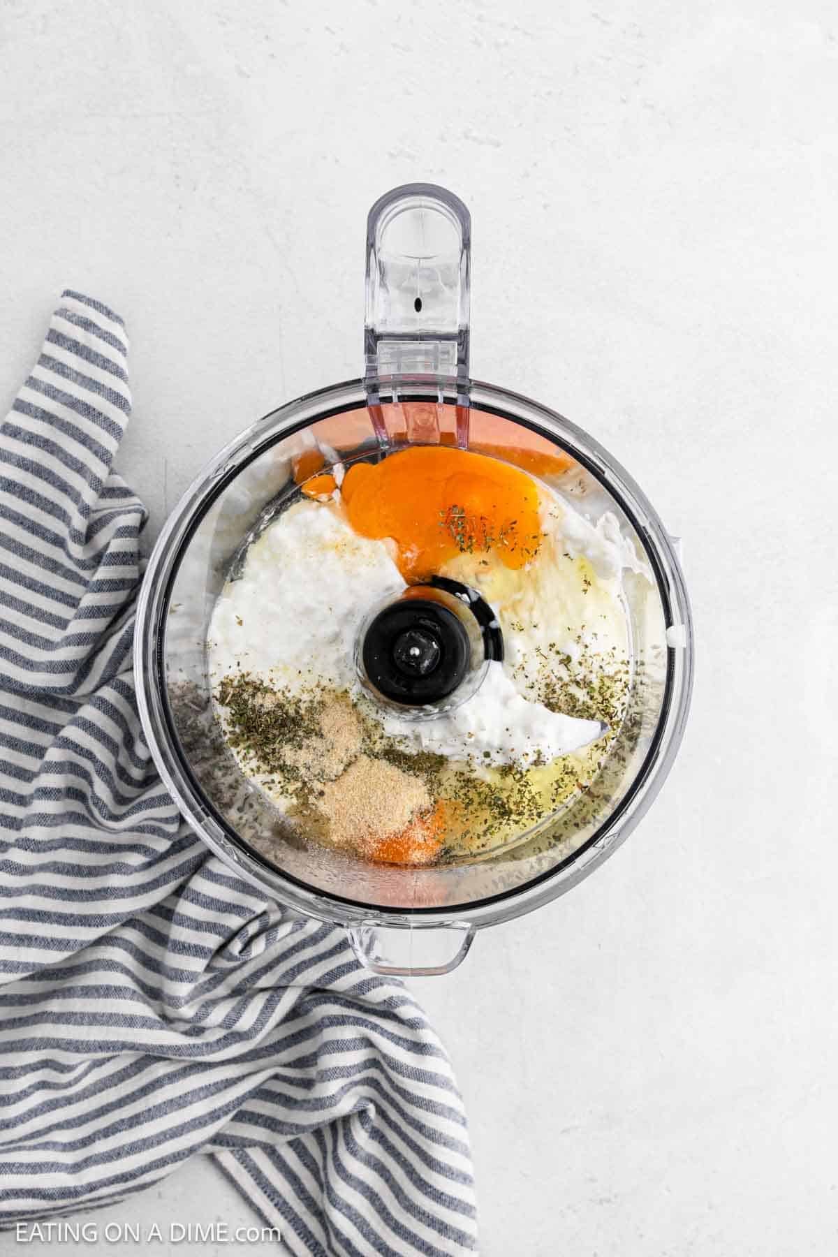 A food processor bowl with eggs, cottage cheese, spices, and herbs inside—perfect for making flatbread—sits on a light surface next to a blue and white striped kitchen towel.