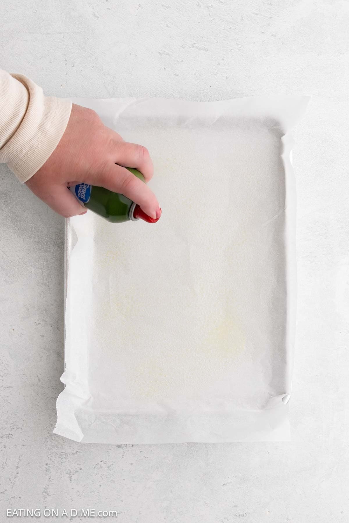 A hand is spraying nonstick cooking spray onto a baking sheet lined with parchment paper, preparing it for baking delicious Low Carb Flatbread. The scene is set on a light-colored countertop.