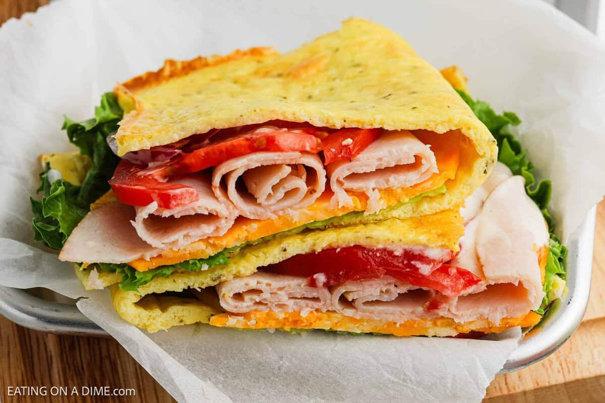 A sandwich with rolled turkey slices, cheddar cheese, lettuce, and tomato, between two folded Cottage Cheese Flatbreads, served on parchment paper.