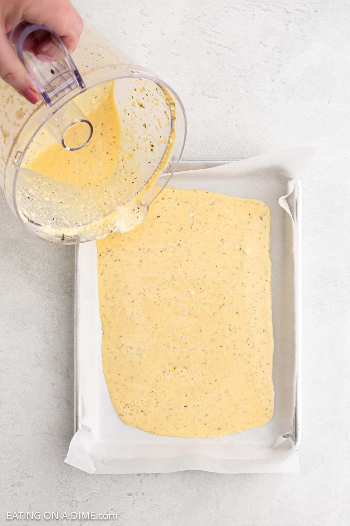 A hand pours a thick, yellow Cottage Cheese Flatbread batter from a food processor onto a parchment-lined baking sheet. The batter spreads out evenly, ready to transform into delicious, protein-packed flatbread.