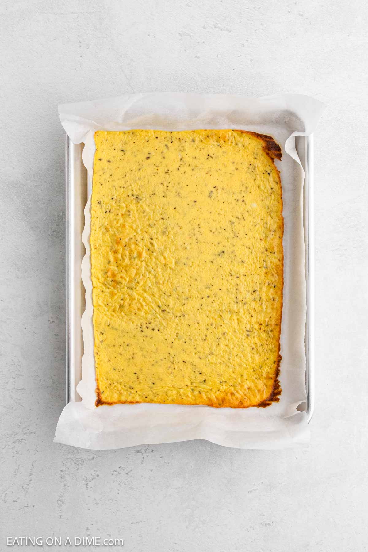 A rectangular, baked yellow flatbread with specks of seasoning sits on parchment paper in a metal baking tray, placed on a light gray surface.