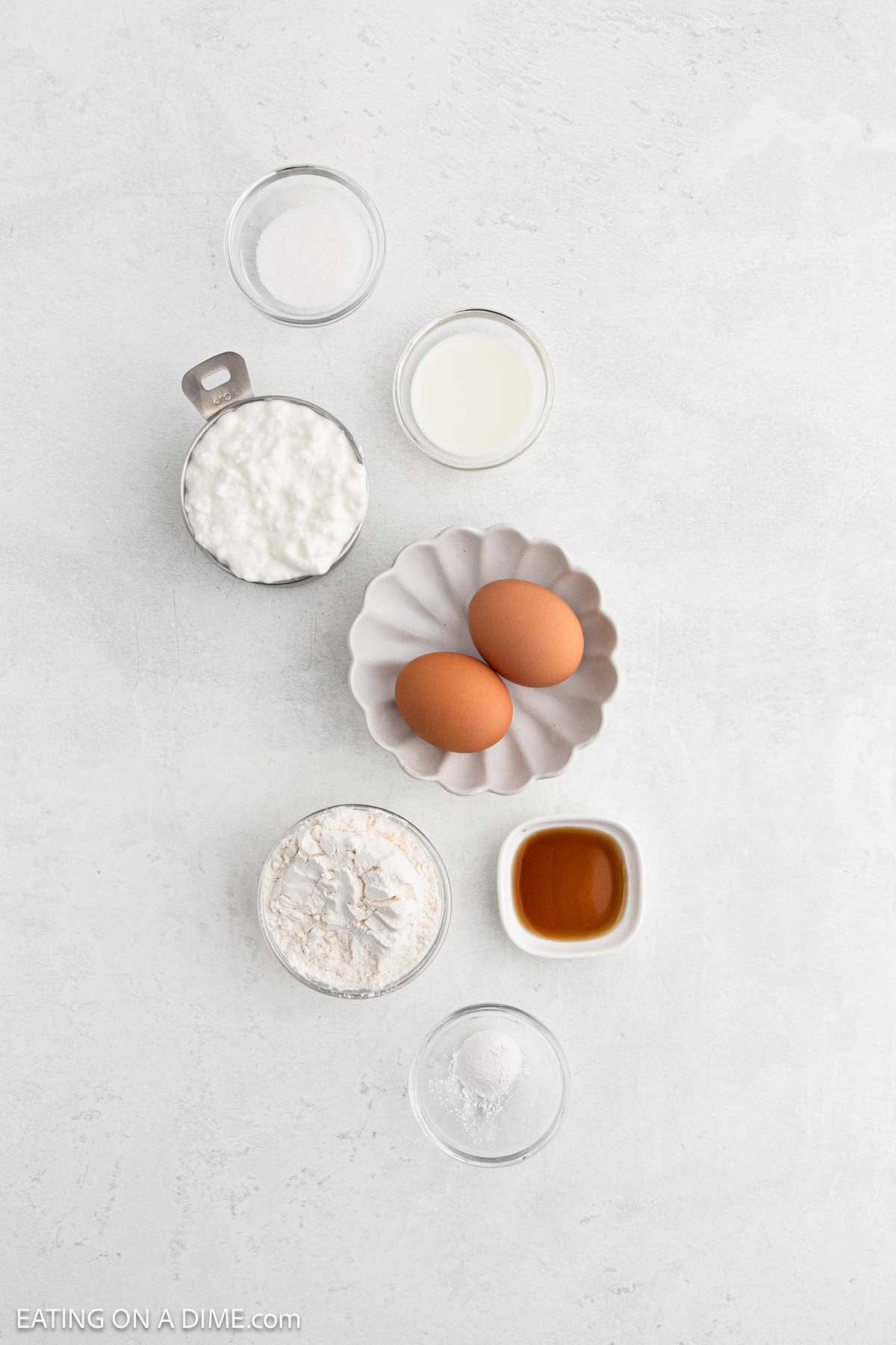 A flat lay of baking ingredients on a white surface, perfect for Cottage Cheese Pancakes: two eggs in a small dish, cottage cheese, flour, milk, sugar, vanilla extract, and baking powder—everything you need for a delicious pancake recipe.
