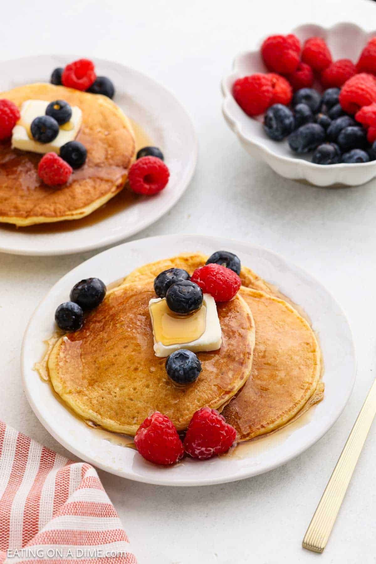 A plate of cottage cheese pancakes topped with butter, blueberries, and raspberries sits on a table next to another stack and a bowl of fresh berries. A red-striped napkin and gold fork complete this healthy pancakes scene.