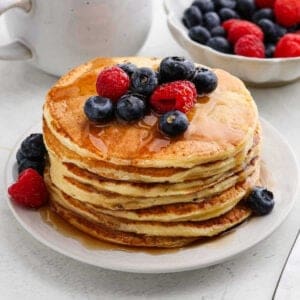 A stack of cottage cheese pancakes topped with fresh blueberries and raspberries, drizzled with syrup, on a white plate. A bowl of berries and a white mug are in the background.