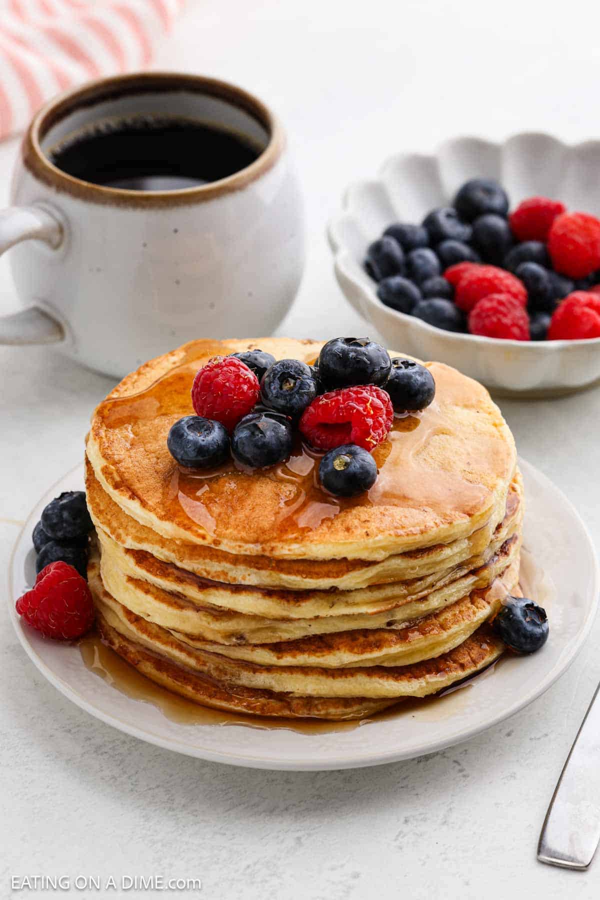 A stack of fluffy cottage cheese pancakes topped with fresh blueberries and raspberries, drizzled with syrup, sits on a white plate. A cup of coffee and a bowl of mixed berries are in the background.