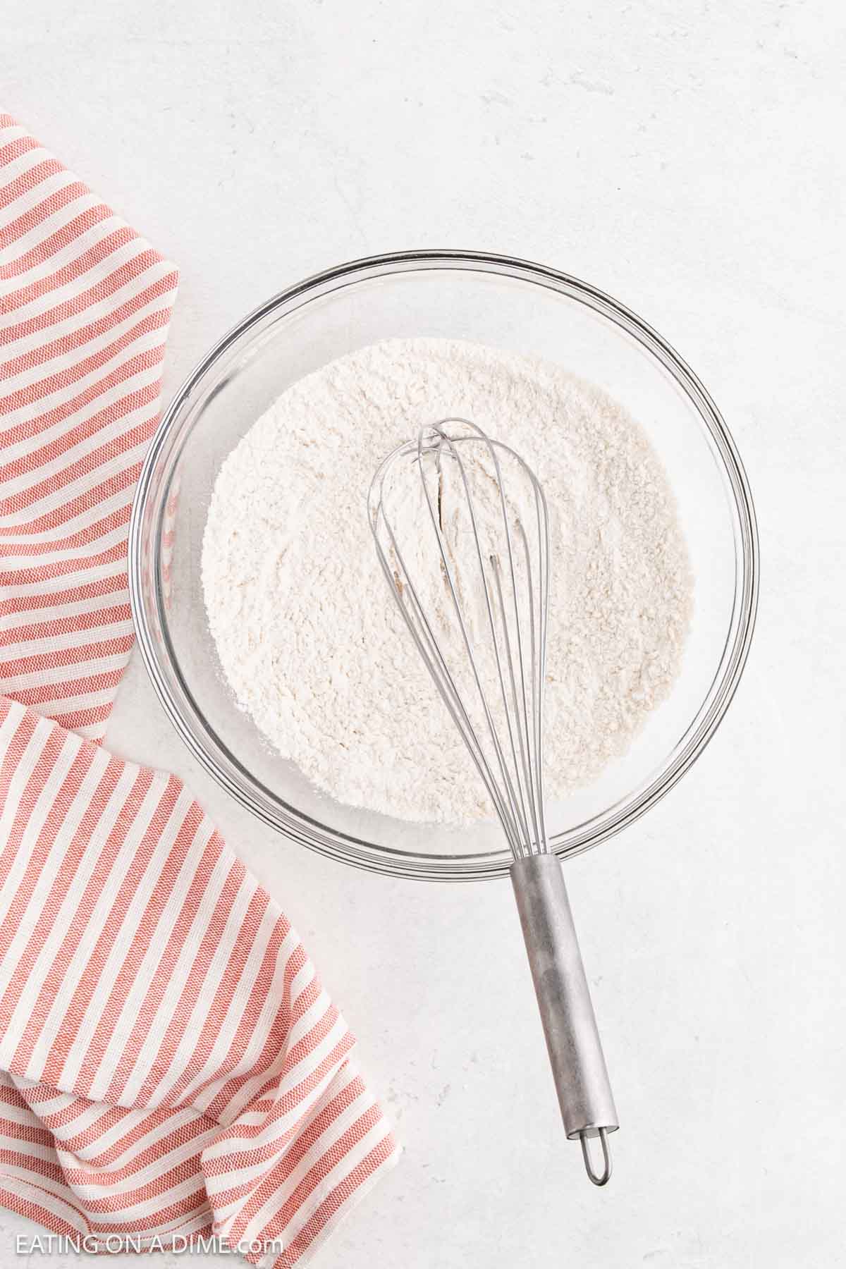A clear glass bowl filled with flour sits on a white surface next to a metal whisk and a red and white striped kitchen towel—perfect for preparing batter for delicious cottage cheese pancakes.