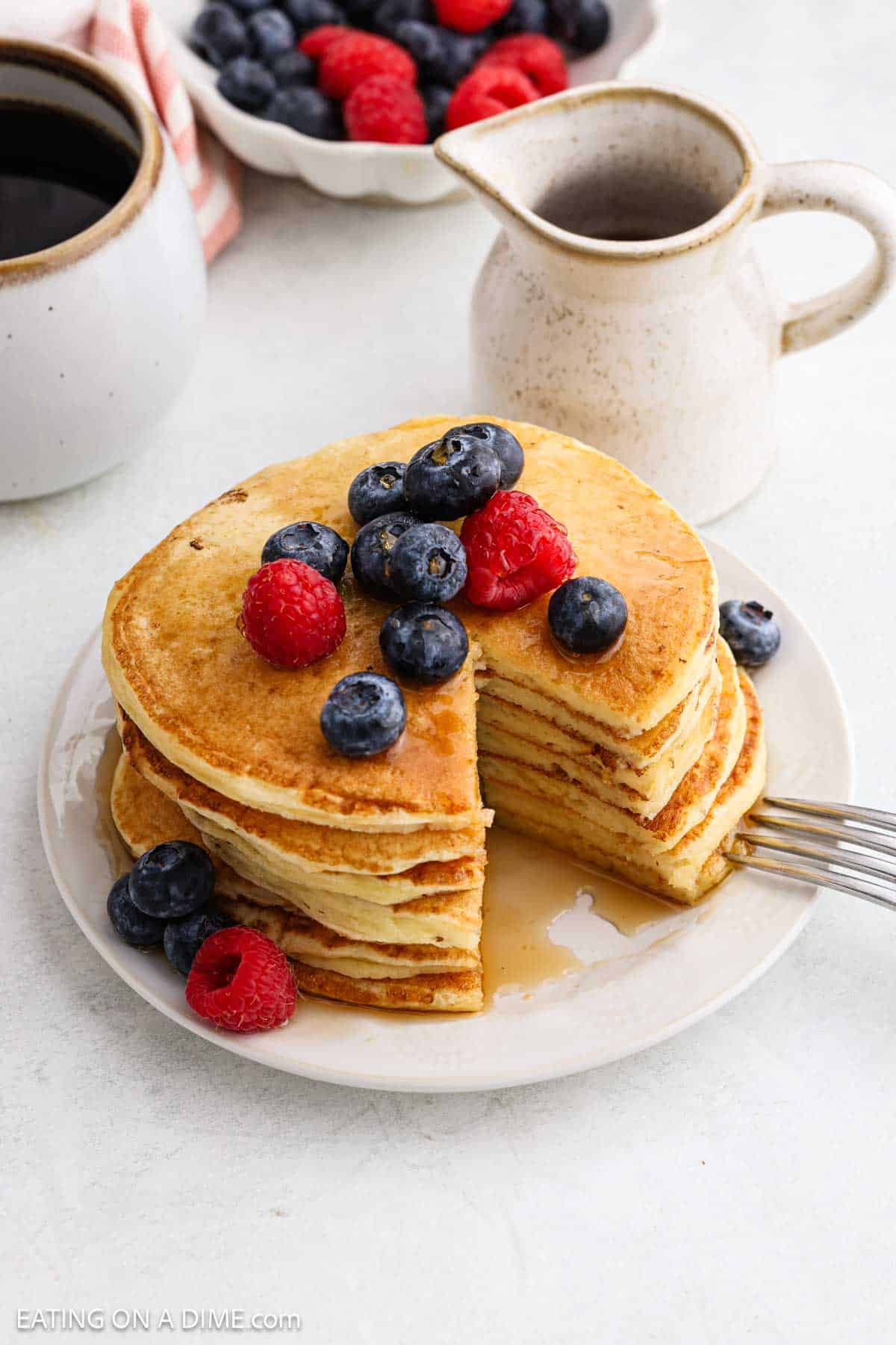 A stack of healthy pancakes, topped with blueberries and raspberries, has a section cut out and syrup poured over. A fork rests on the plate, while a cup, a small jug, and a bowl of berries complete this delicious cottage cheese pancakes scene.