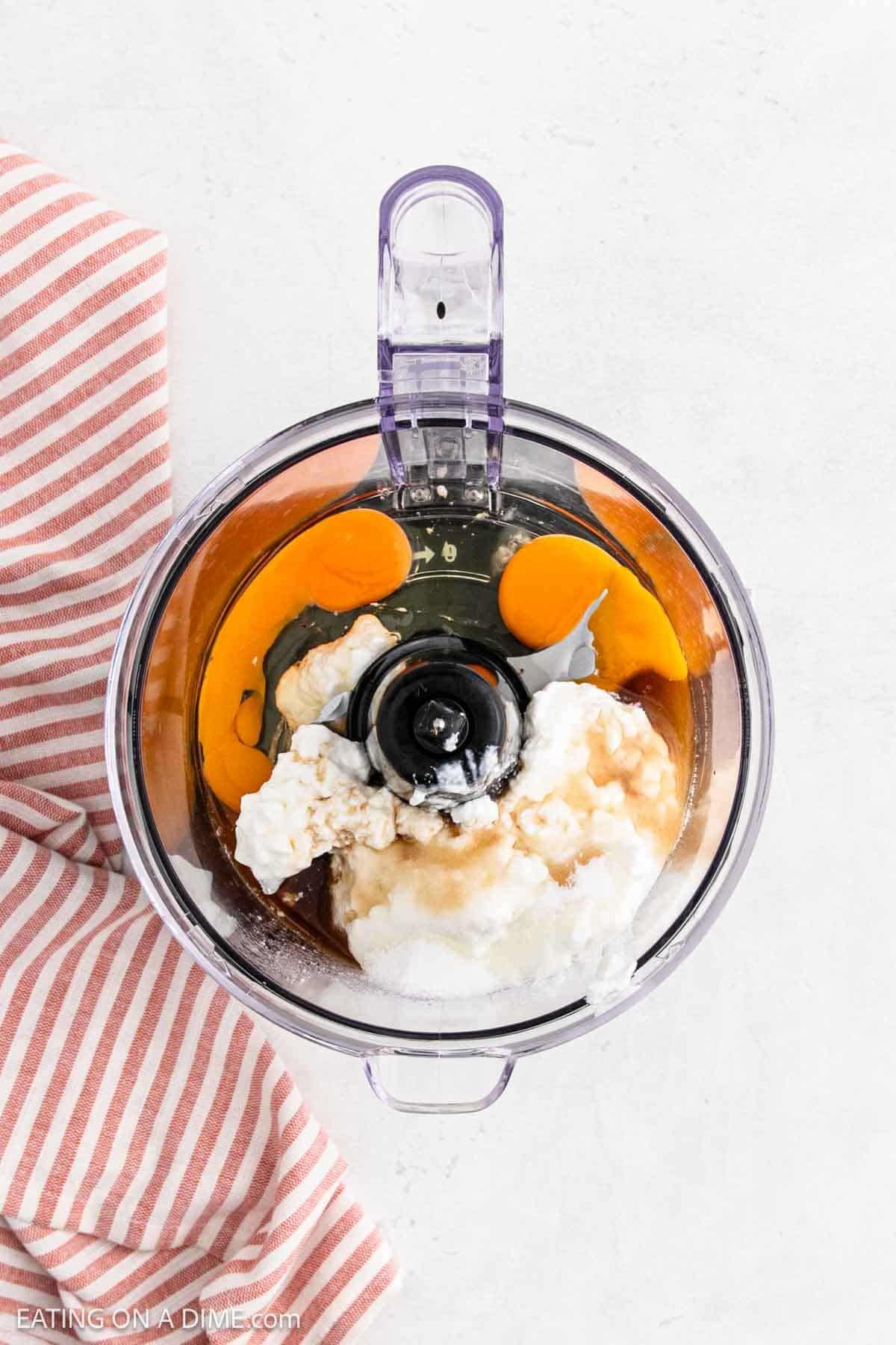 A food processor bowl with eggs, cottage cheese, sugar, and vanilla extract inside—perfect for making cottage cheese pancakes—is viewed from above. A red and white striped kitchen towel rests beside the bowl on a white surface.