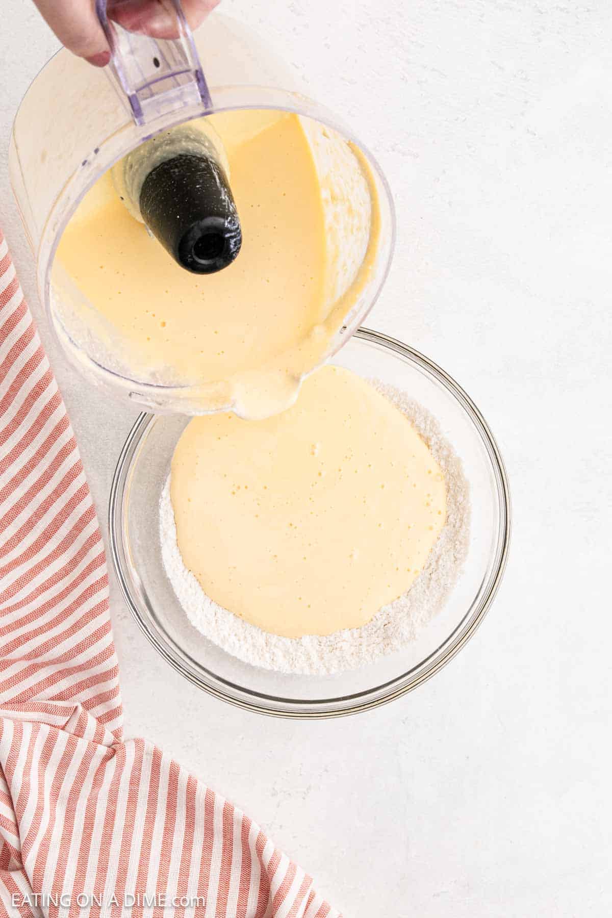 A glass bowl of flour on a white surface, with yellow batter for a Cottage Cheese Pancakes recipe being poured in from a food processor. A pink and white striped cloth sits beside the bowl.