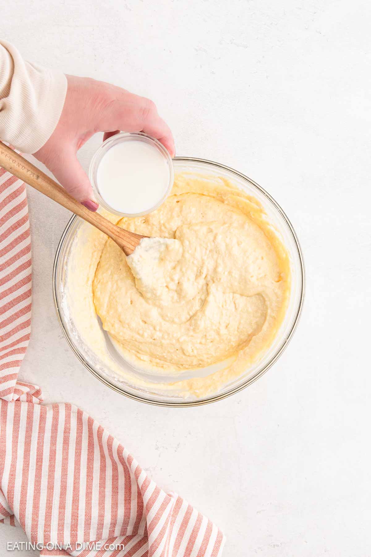 A hand holds a small glass cup of milk above a clear bowl filled with yellow cottage cheese pancake batter, being stirred with a wooden spoon. A pink and white striped cloth rests nearby on the white surface.