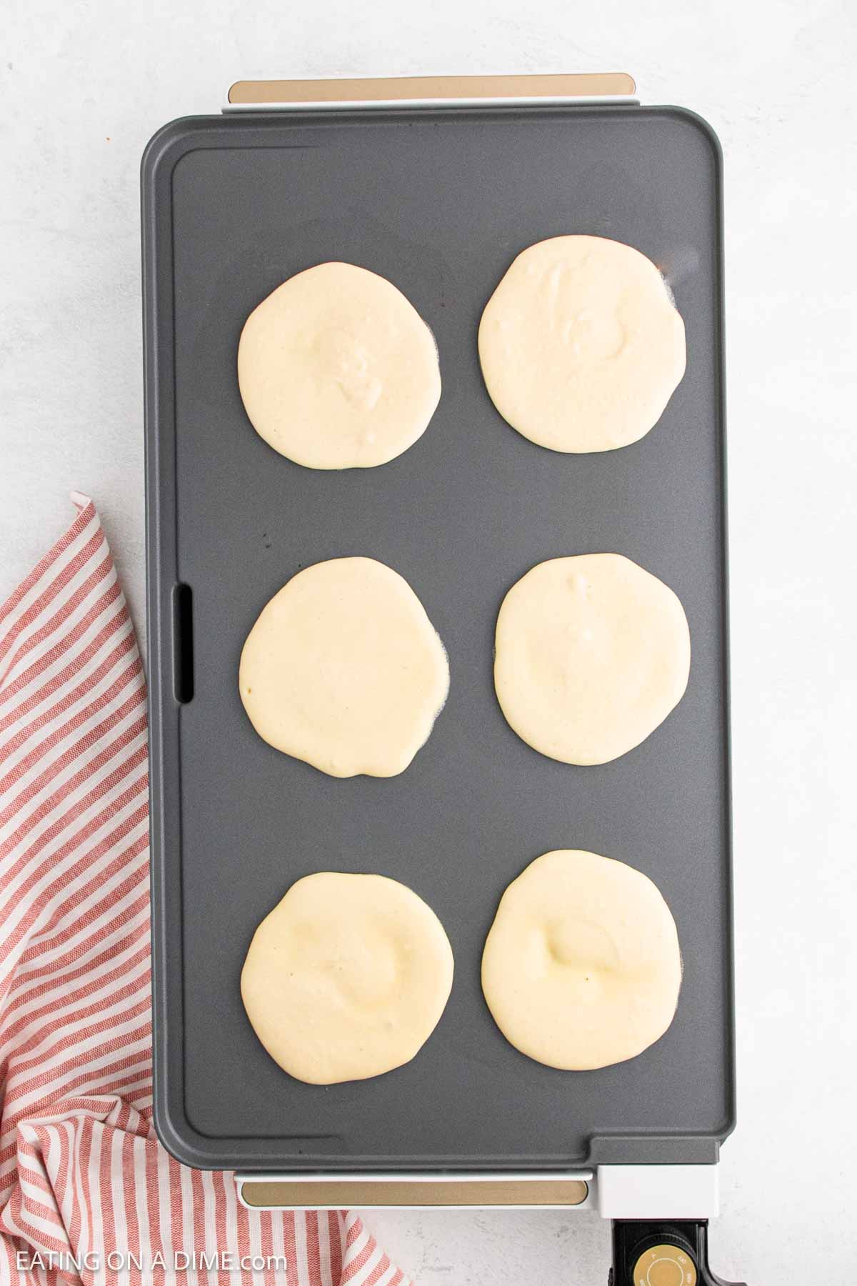 An electric griddle with six cottage cheese pancakes cooking on it, next to a red and white striped kitchen towel on a light surface.