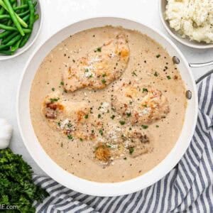 A skillet with Creamy Garlic Chicken breasts garnished with herbs, surrounded by bowls of green beans, mashed potatoes, grated cheese, garlic bulbs, and fresh parsley on a striped cloth.