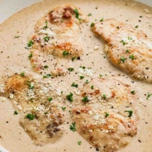 Four pieces of Creamy Garlic Chicken breast in a rich white sauce, garnished with chopped parsley, grated cheese, and cracked black pepper on a plate.