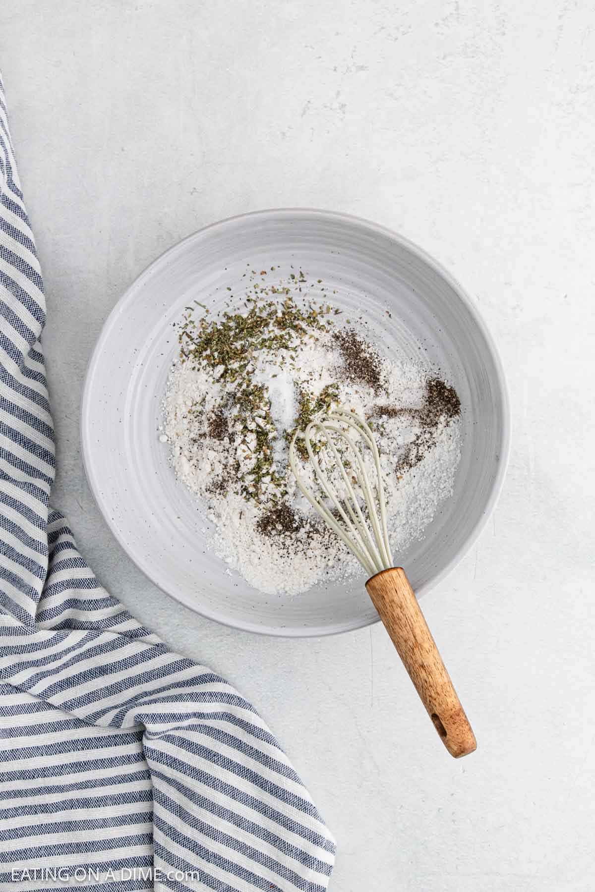 A white bowl with flour and seasonings being whisked together, perfect for making Creamy Garlic Chicken, is placed on a light surface next to a blue and white striped kitchen towel.