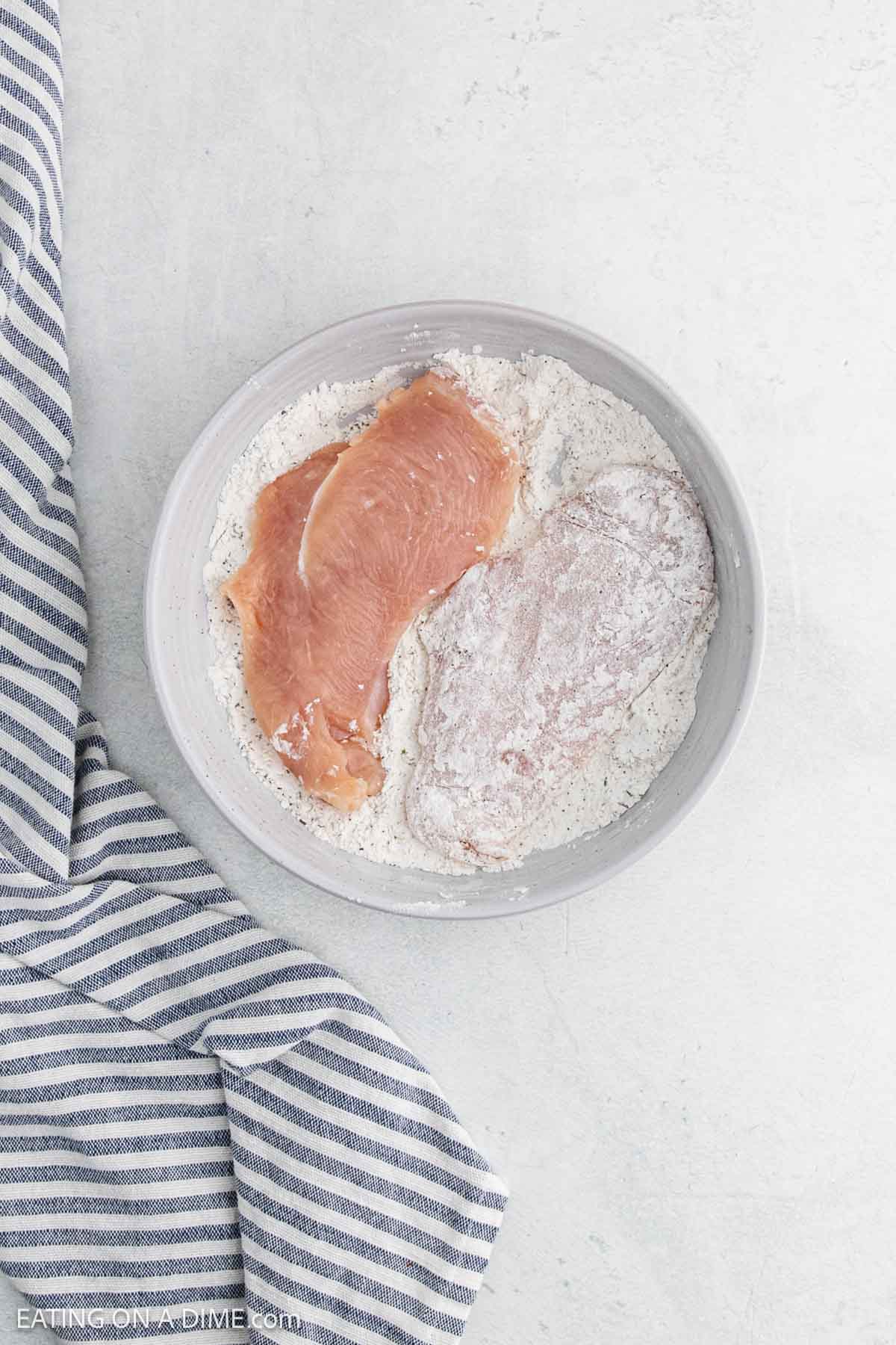 A bowl with flour and seasonings holds two raw chicken breast fillets—one coated, one uncoated—ready for creamy garlic chicken. A blue and white striped kitchen towel rests beside the bowl on a light surface.